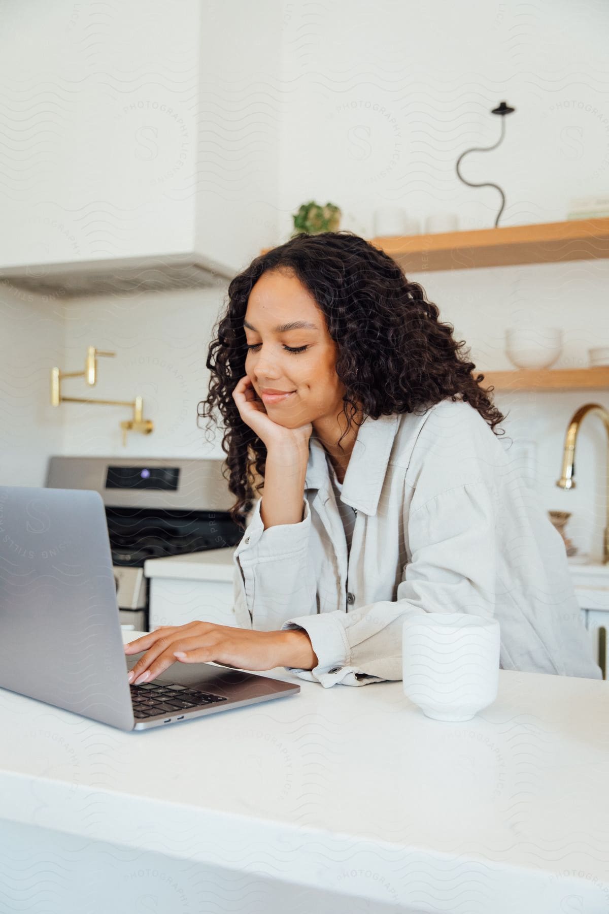 A smiling woman sitting at a laptop on a kitchen counter.