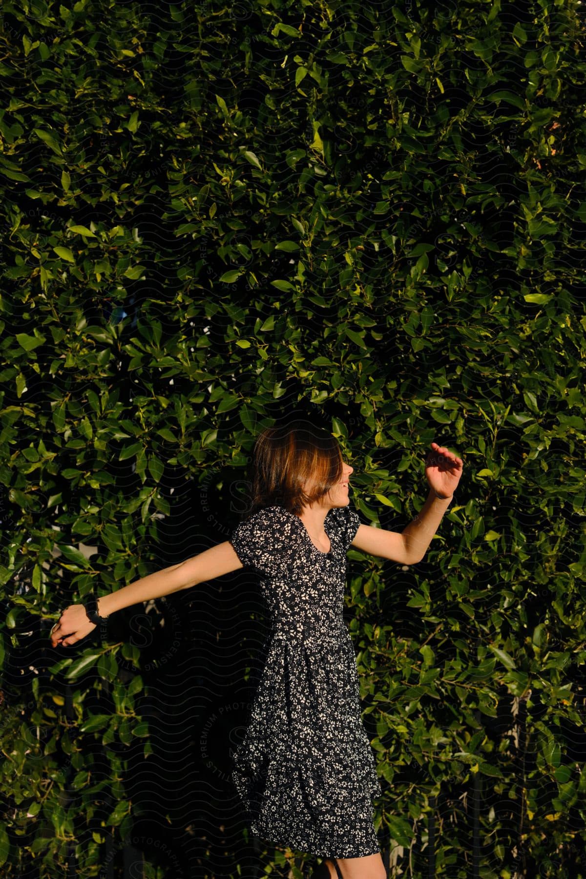 Brunette woman in floral dress stands before a tall green hedge.