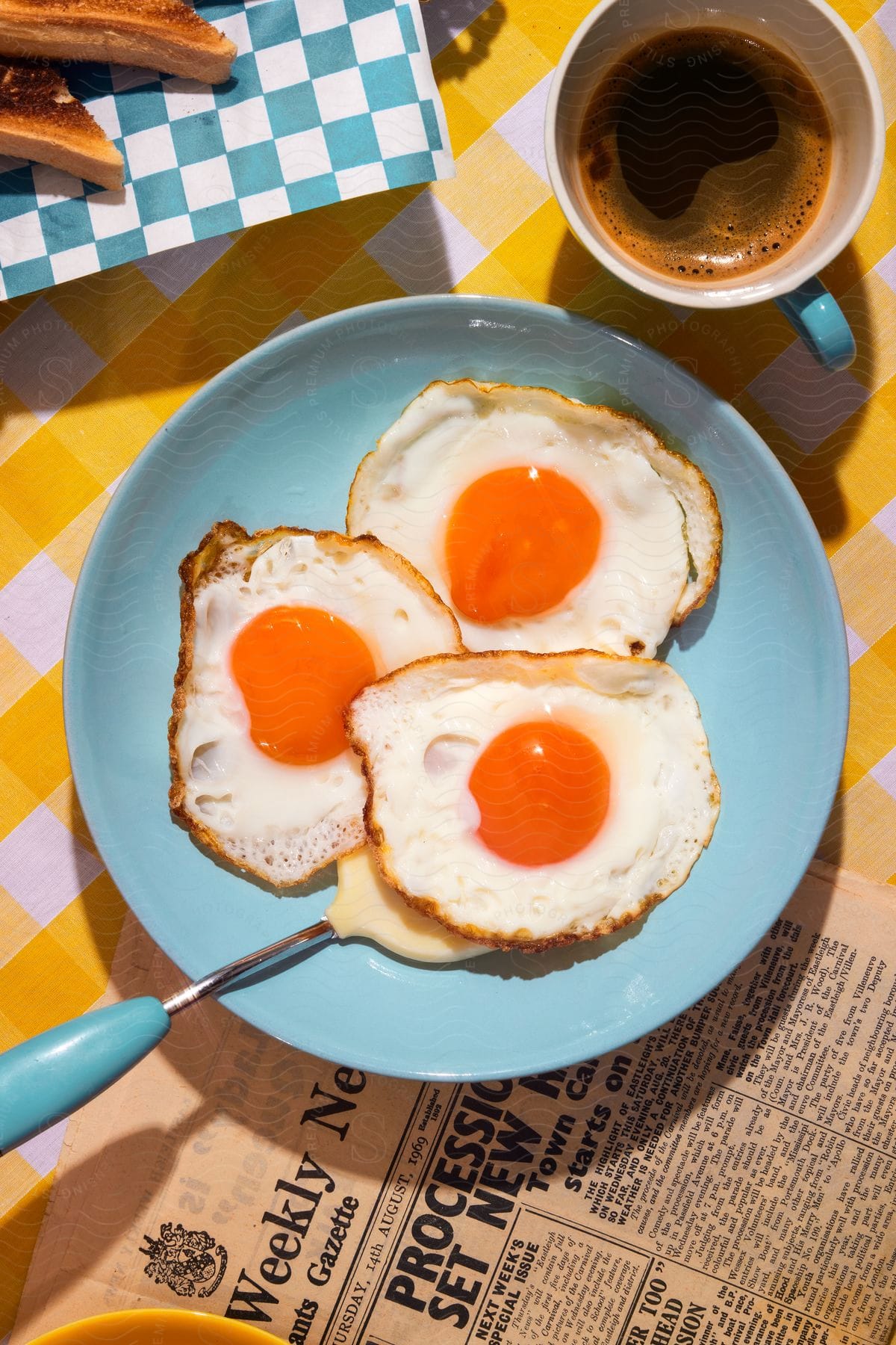 Three sunny-side up eggs on a blue plate with a blue and white checkered napkin with toast and a cup of black coffee.