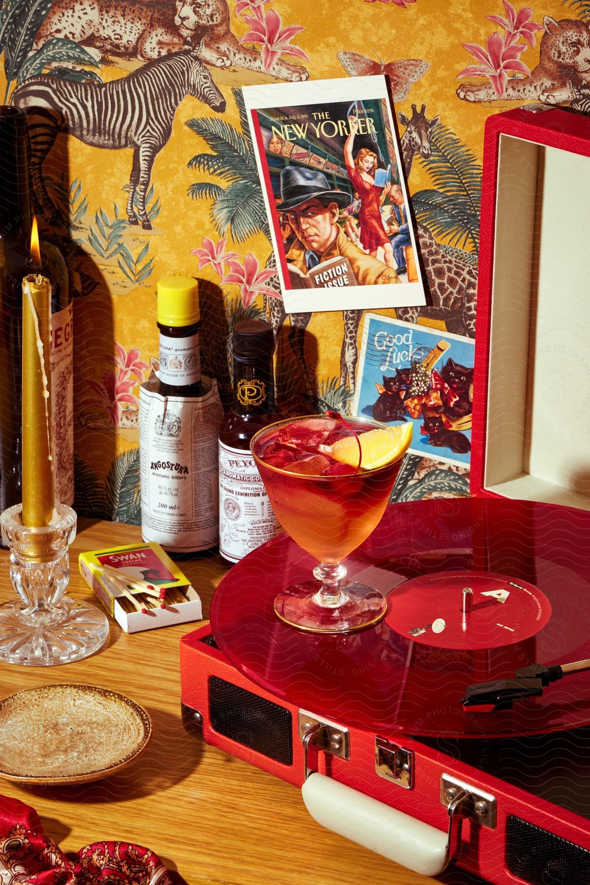 A cocktail sits on a red vinyl record on a turntable with a candle and bottles of alcohol on the table