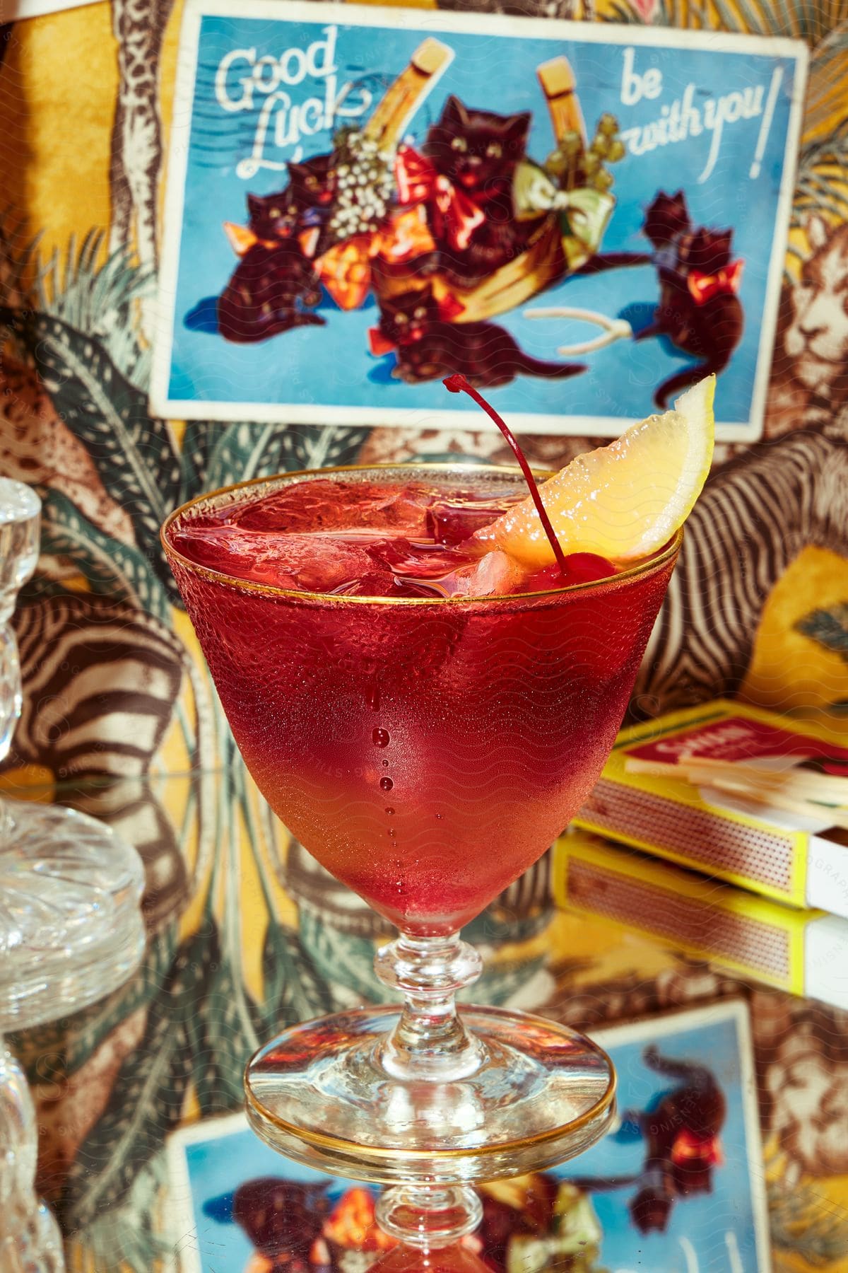 A cocktail garnished with lemon wedge and a cherry set on a glass table under a postcard and colorful decor