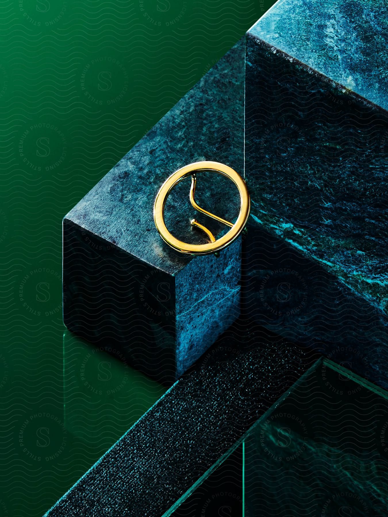 Gold circular brooch on dark green marble block with reflective surface and contrasting textures.