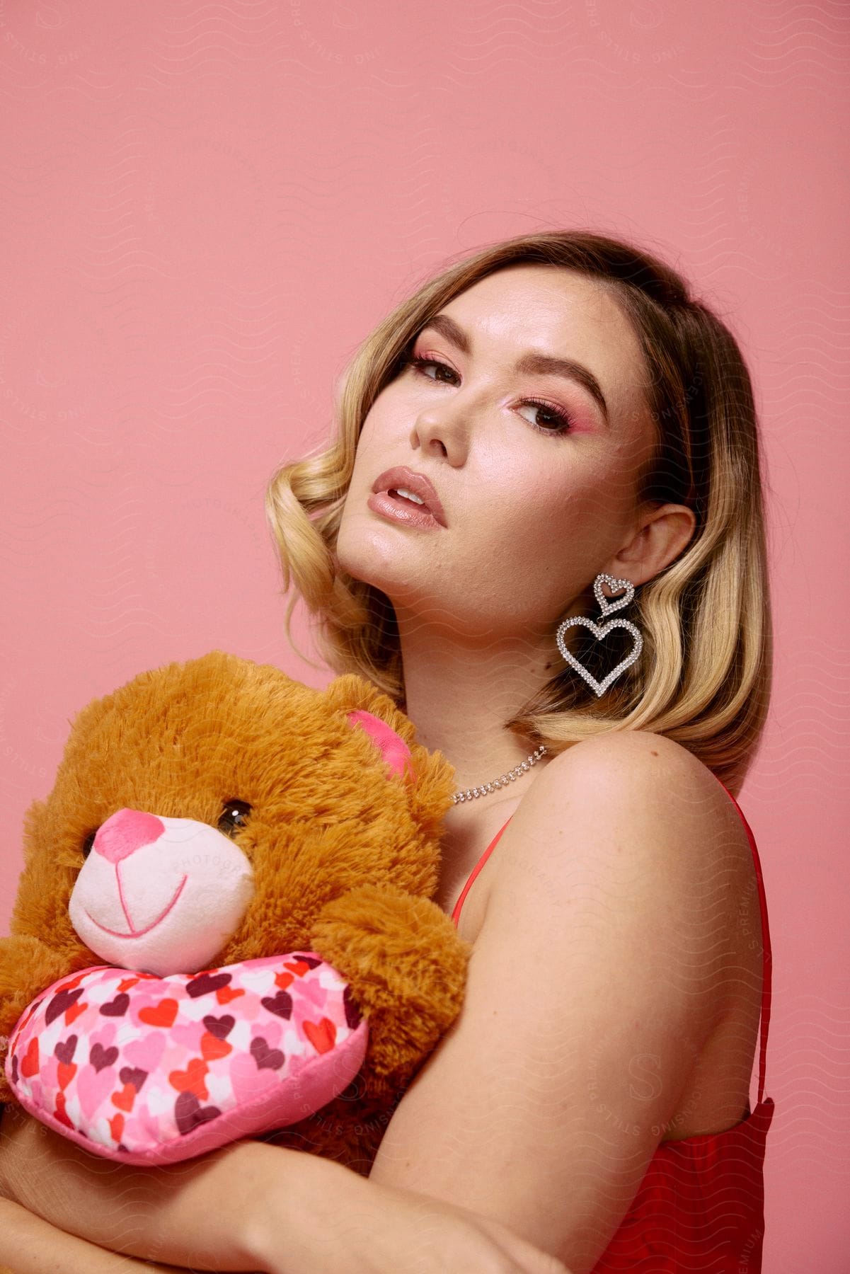 A beautiful woman holding a teddy bear with matching heart accessories in a pink background