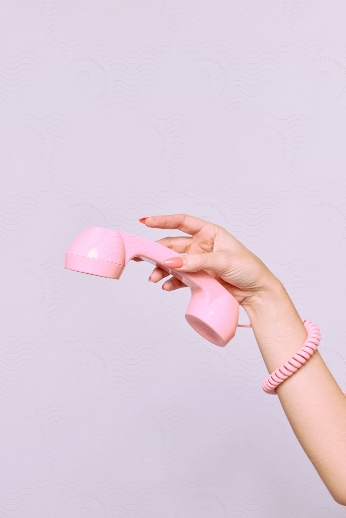 A woman holds a pink rotary phone receiver in her hand with the cord wrapped around her wrist.