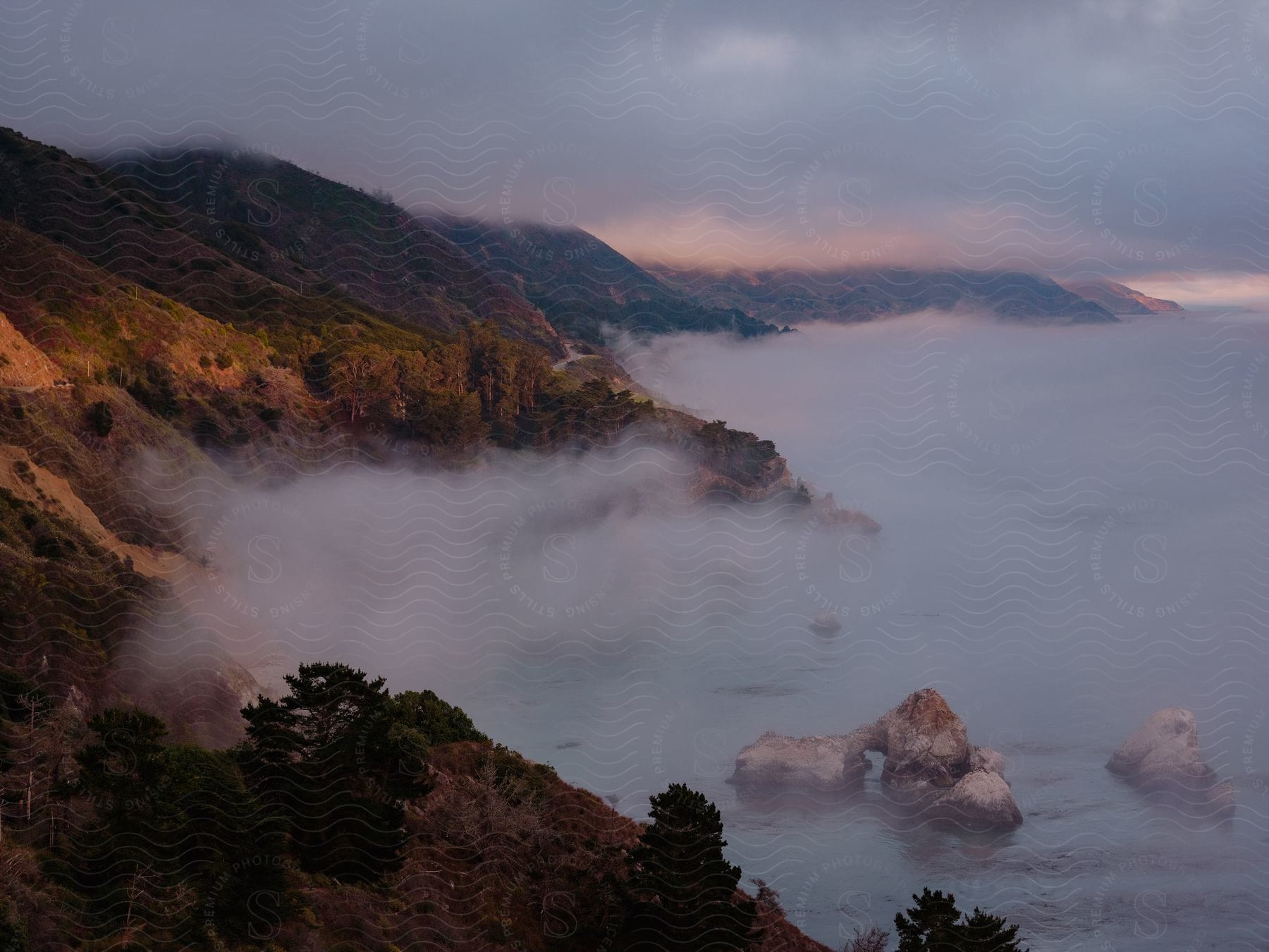 A fog layer at the base of oceanside cliffs and hills with heavy cloud cover above