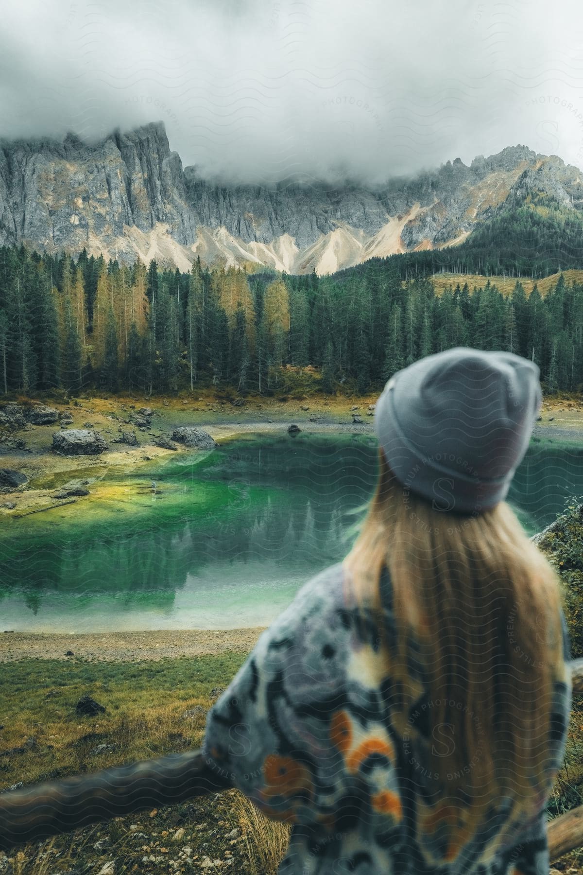 A person from the back, wearing a gray hat and a colorfully patterned jacket, looking at a mountainous landscape and a lake.