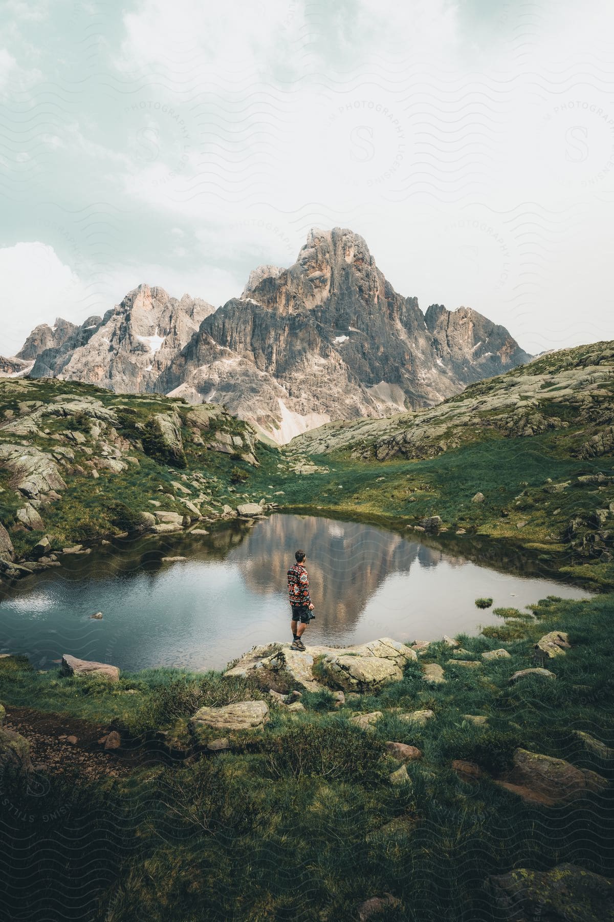 A person stands in front of a lake that is reflecting a mountain with rugged peaks in the background.