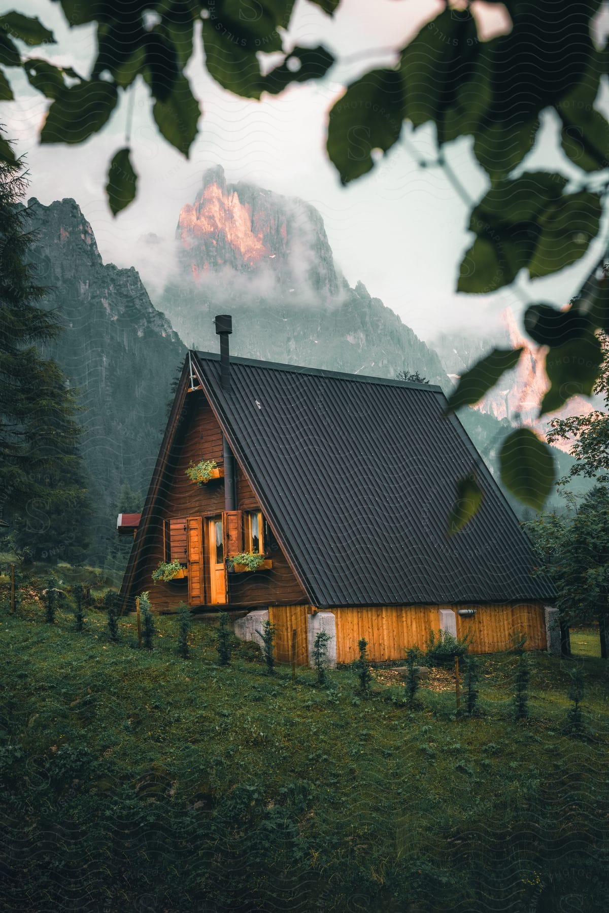 An A-frame house sits on a hillside beneath a steep mountain range in the summertime.