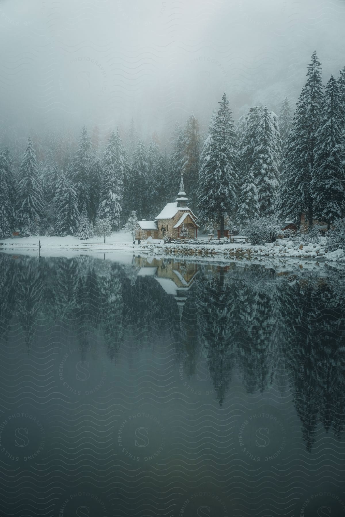 Still waters reflect snow covered church and conifer forest on a foggy day.