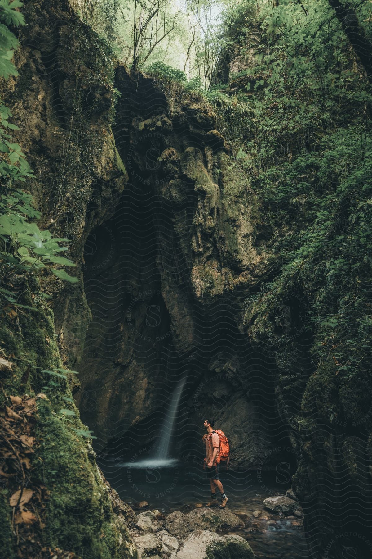 Hiker stands near a waterfall at the foot of a cliff holding the strap of his backpack