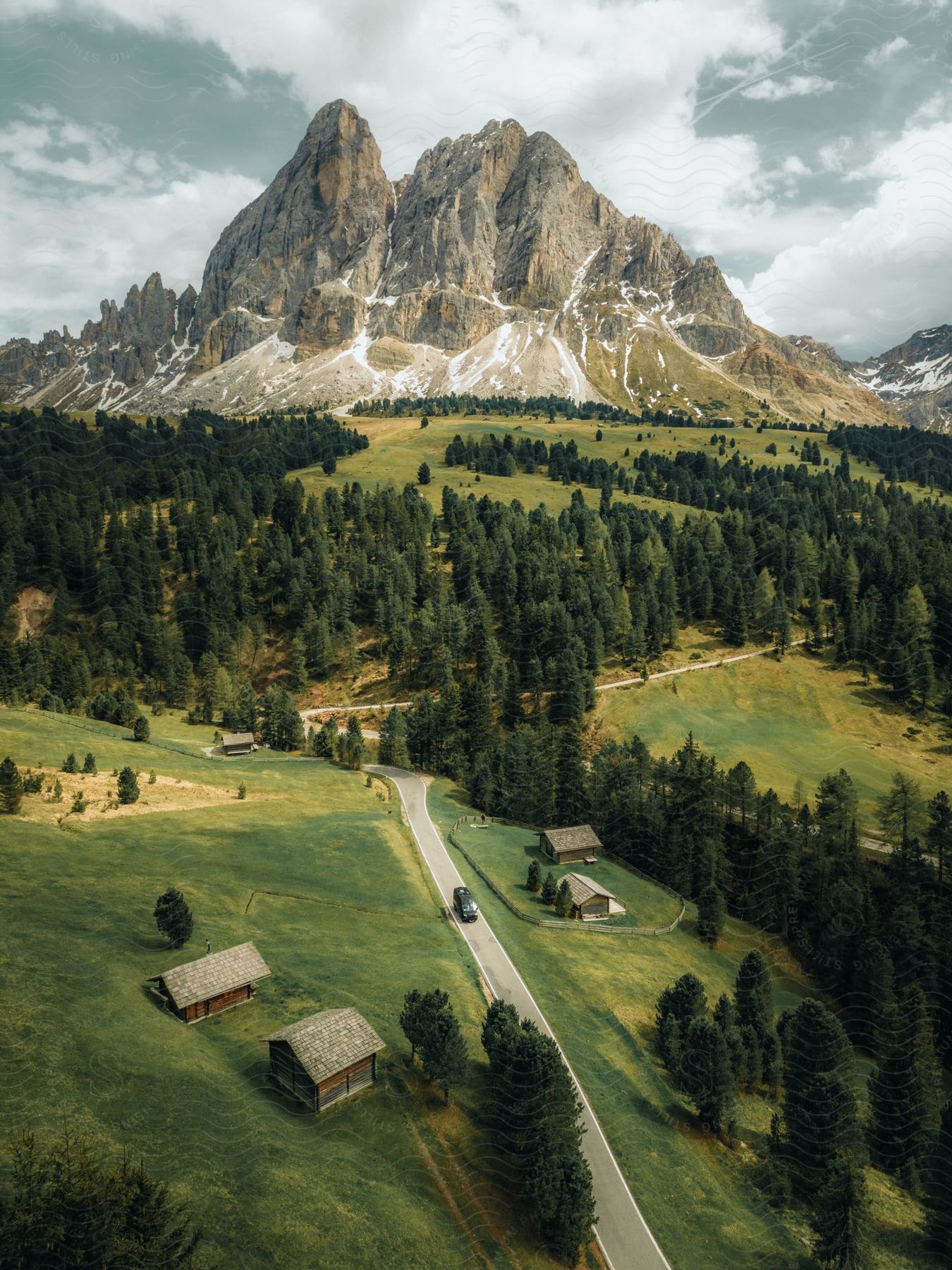 A winding road leading through a valley with sparse wooden huts and a backdrop of towering, craggy mountain peaks.