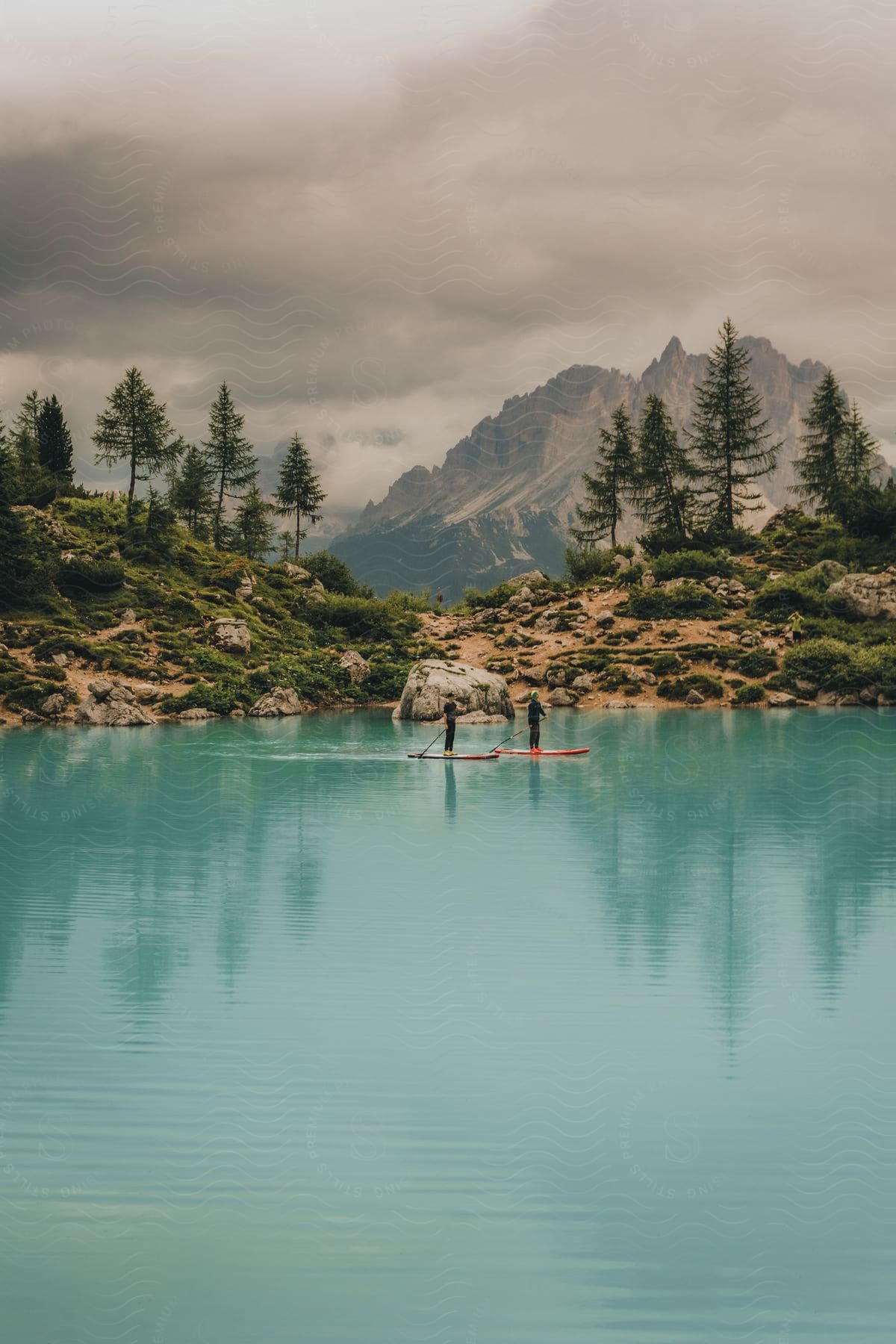 A turquoise lake surrounded by rocky terrain and green pine trees, with two people paddleboarding in the middle of the lake and mountains in the background partially covered by clouds.