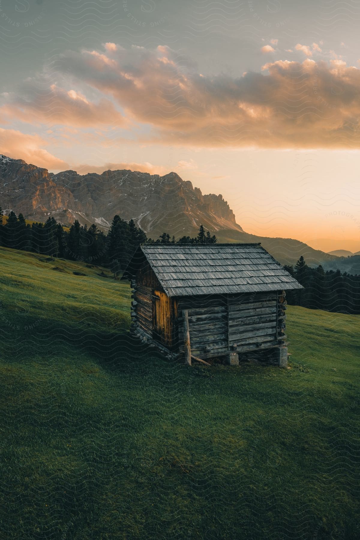 Small wooden cabin set in a lush green countryside with majestic mountains and a sunset sky as a backdrop.