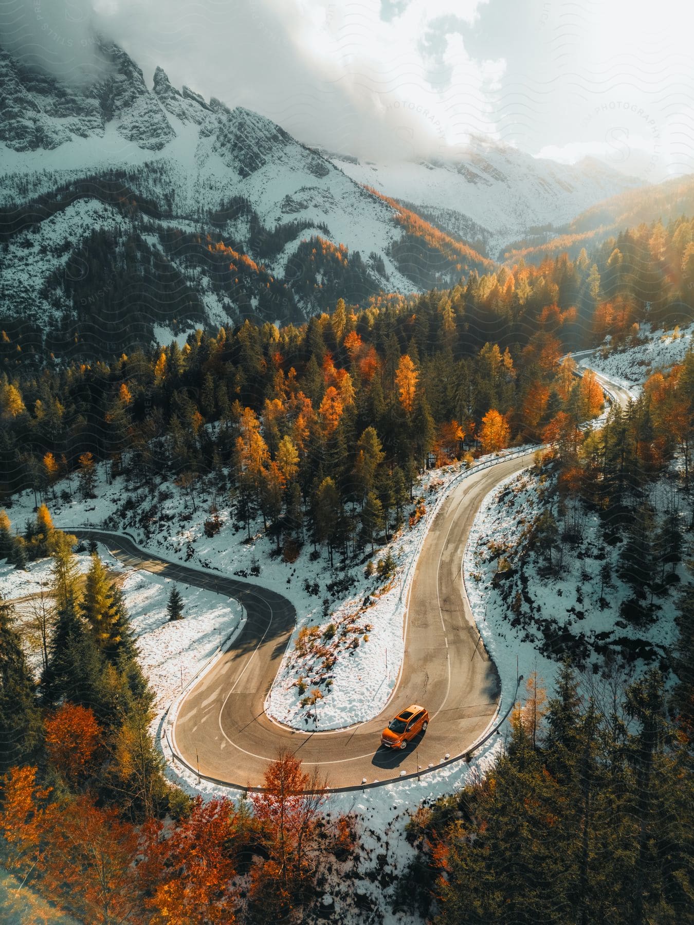 A view of a curved road amidst a mountainous landscape with snow and pine trees with an orange car driving on the asphalt.