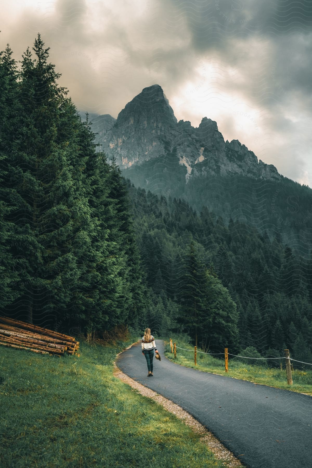 A person walking along an asphalt road that cuts through a green forest and in the background is an imposing and rugged mountain that is partially covered by fog.