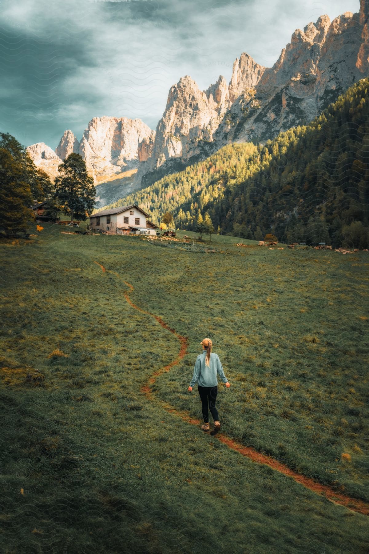 A woman is walking towards a mountainous backdrop, following a narrow dirt path through a green field with a house nearby.