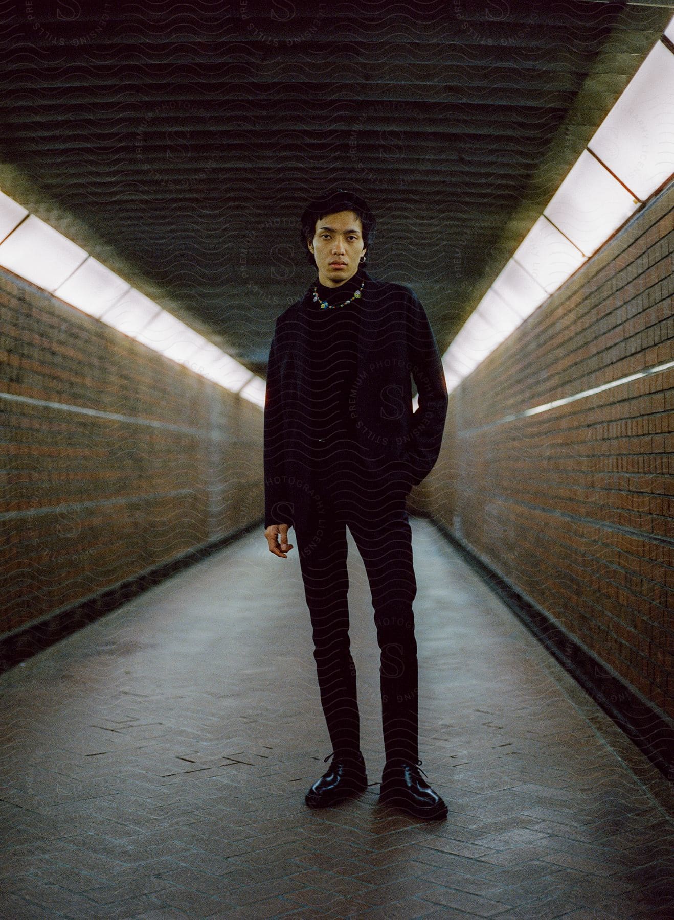 A suited man in black, hands in pockets, stands alone in a tunnel.