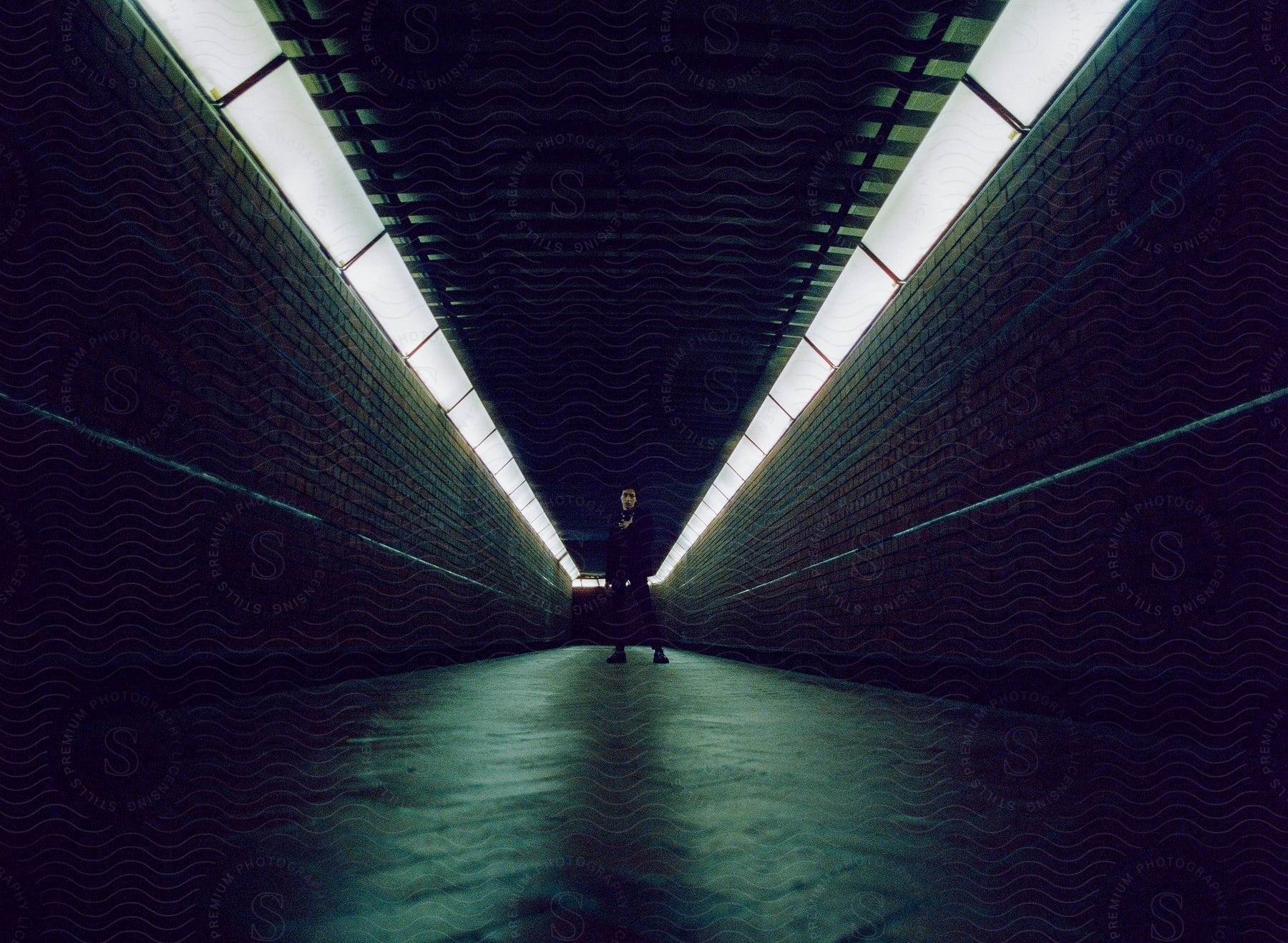 Man standing at the end of a dimly lit tunnel with fluorescent lights and brick walls.
