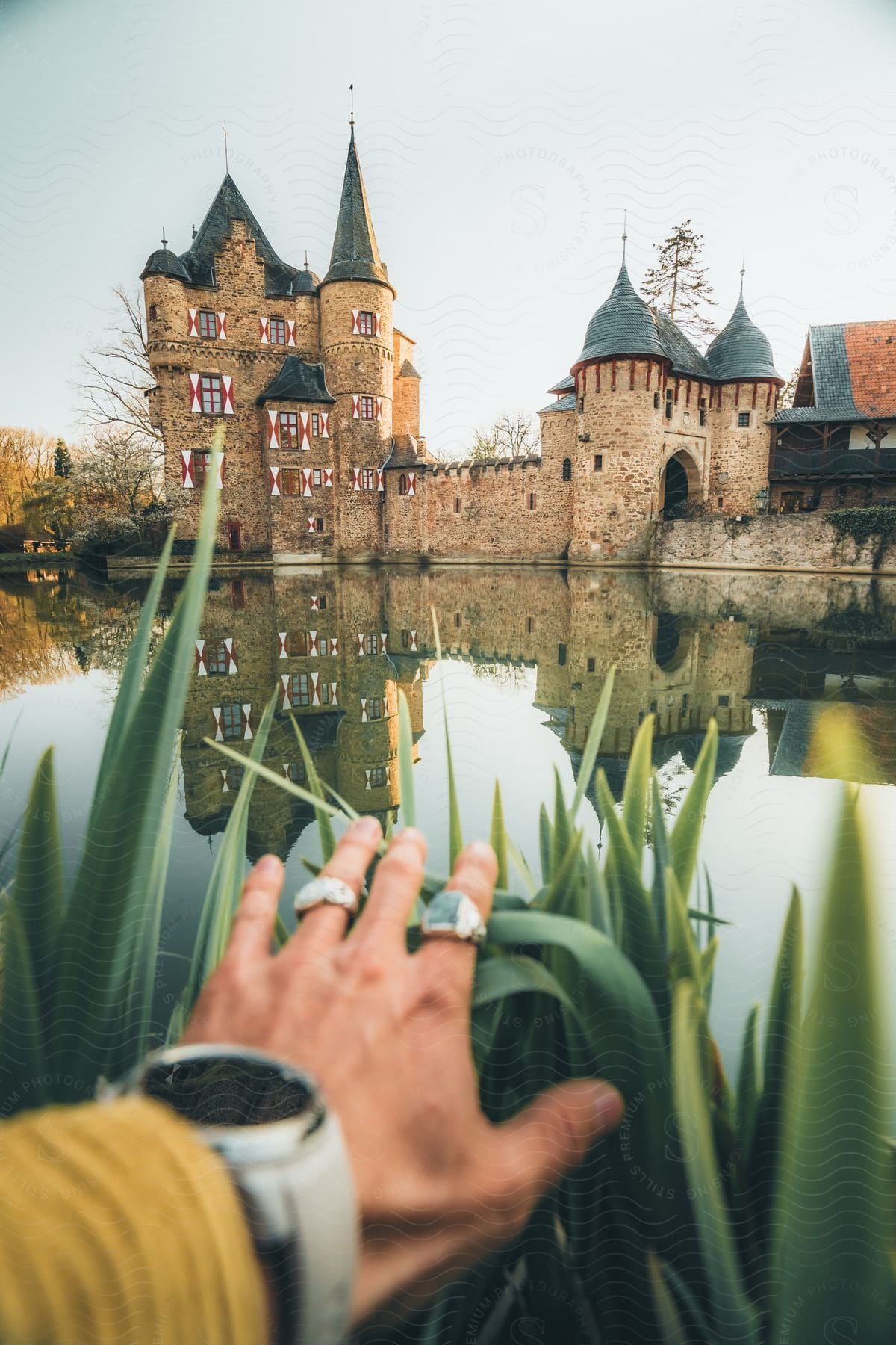 Person with outstretched hand toward moated castle reflecting on water.