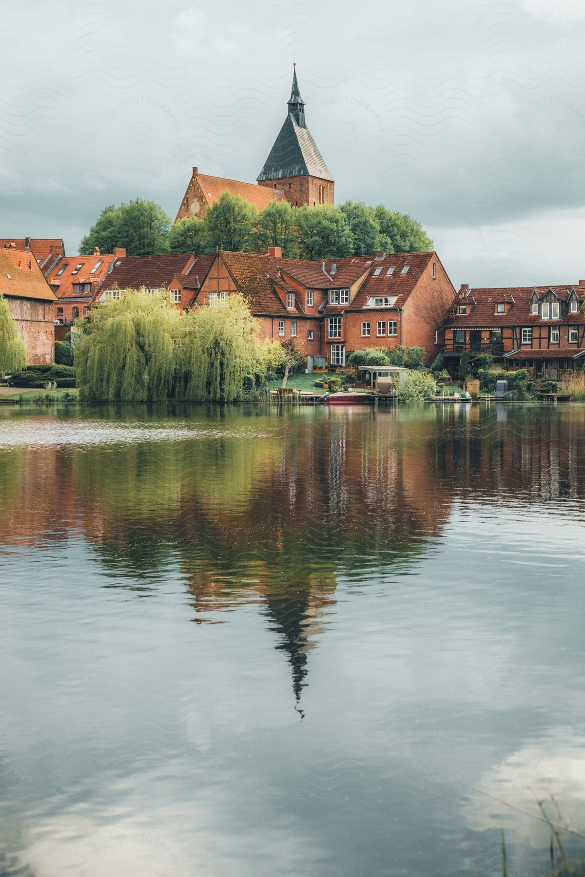 Church spire and reflection on water, framed by trees and residential buildings, on cloudy day.