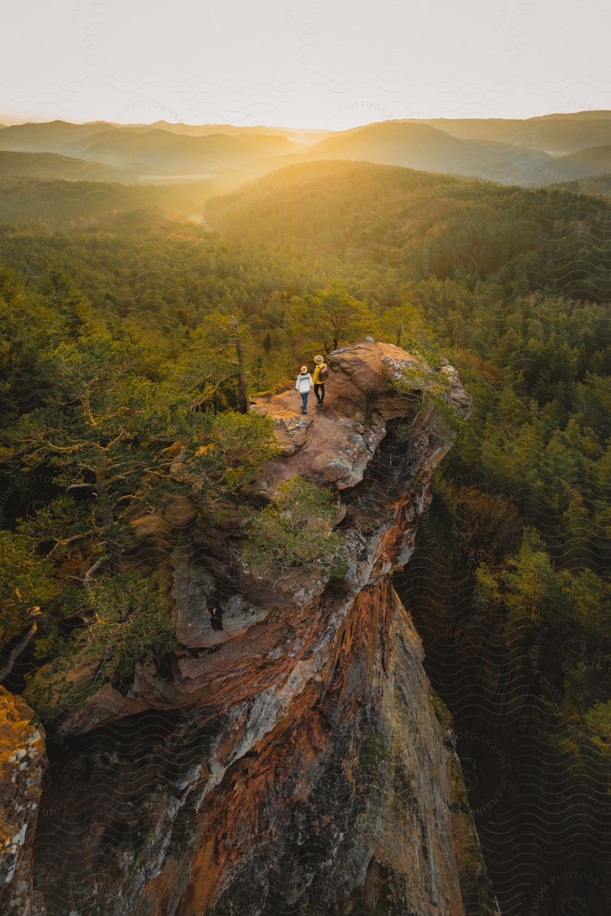 Two people standing on a mountain cliff overlooking forests and mountains as the sun shines on the horizon