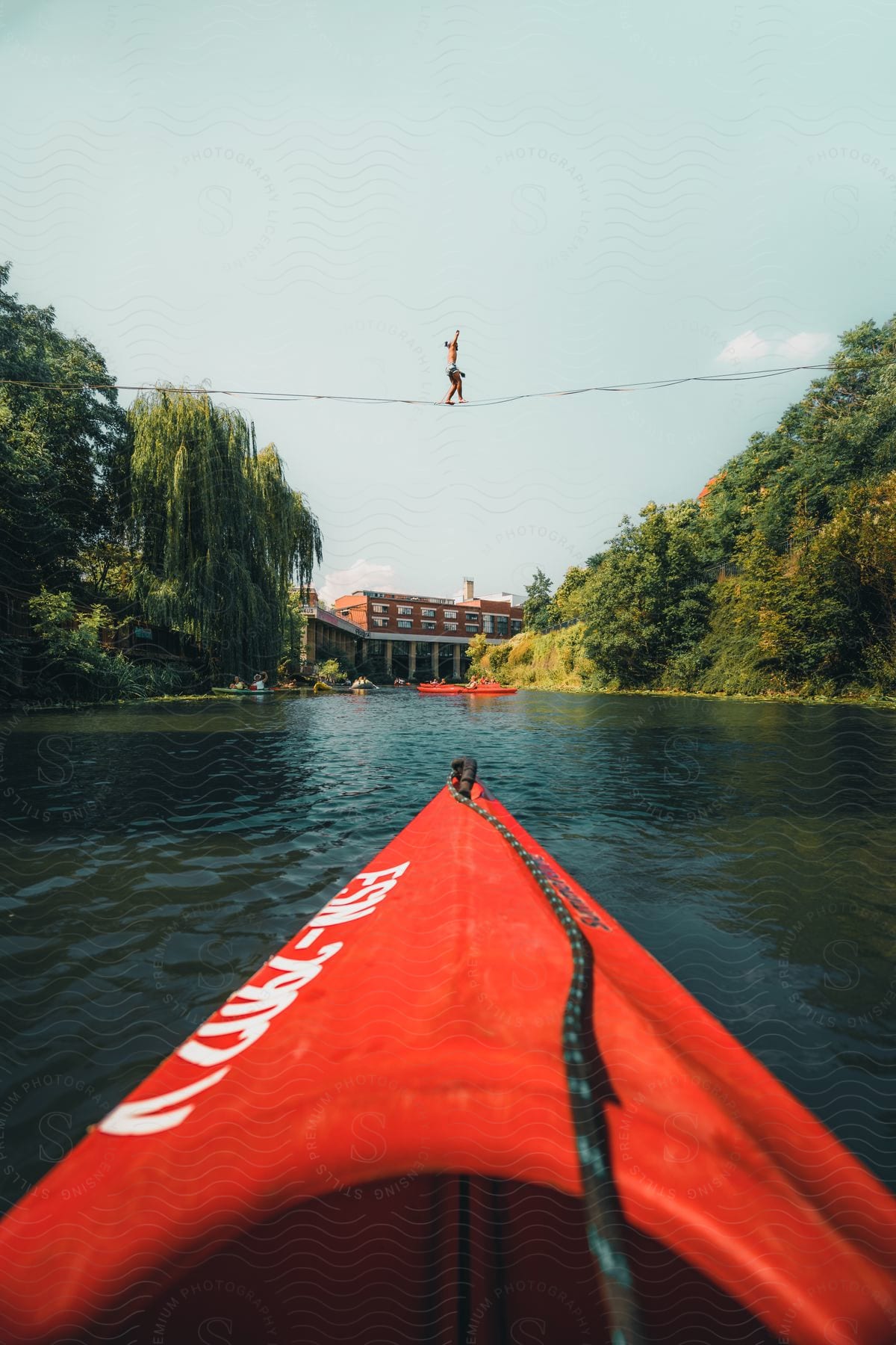 Stock photo of a person balances on a slackline stretched across a river, observed from a red canoe floating nearby.