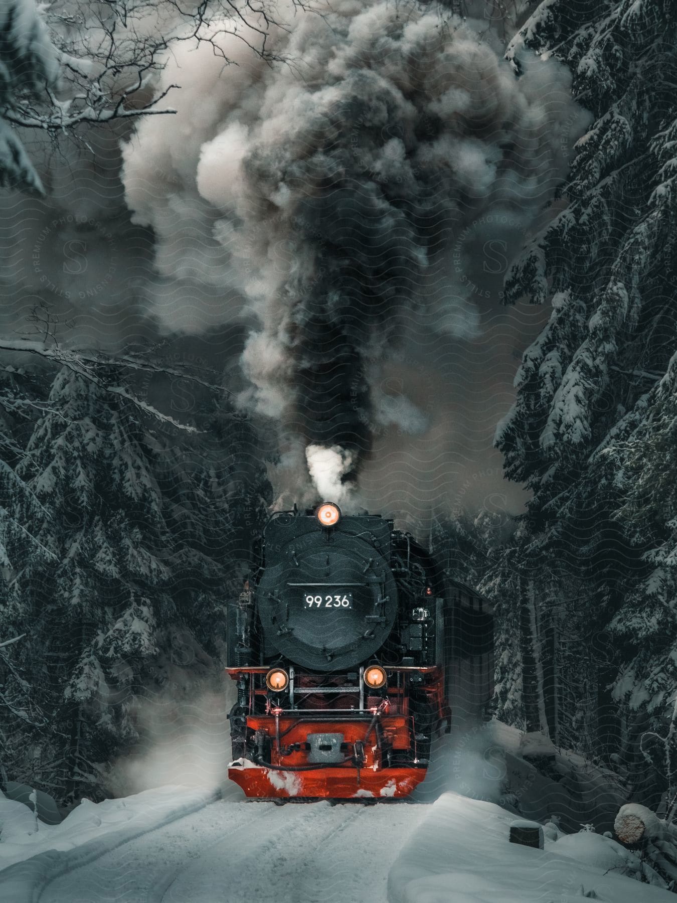 A black and red train driving in a region with snowy ground and some trees on the side and is releasing a lot of smoke and upwards