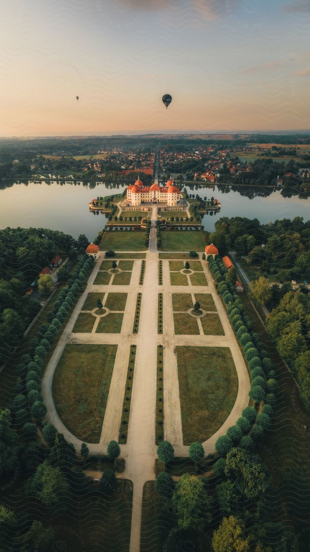 A large and well-kept garden in front of a large mansion surrounded by a lake with lots of vegetation around and a civilization in the background with balloons flying in the sky