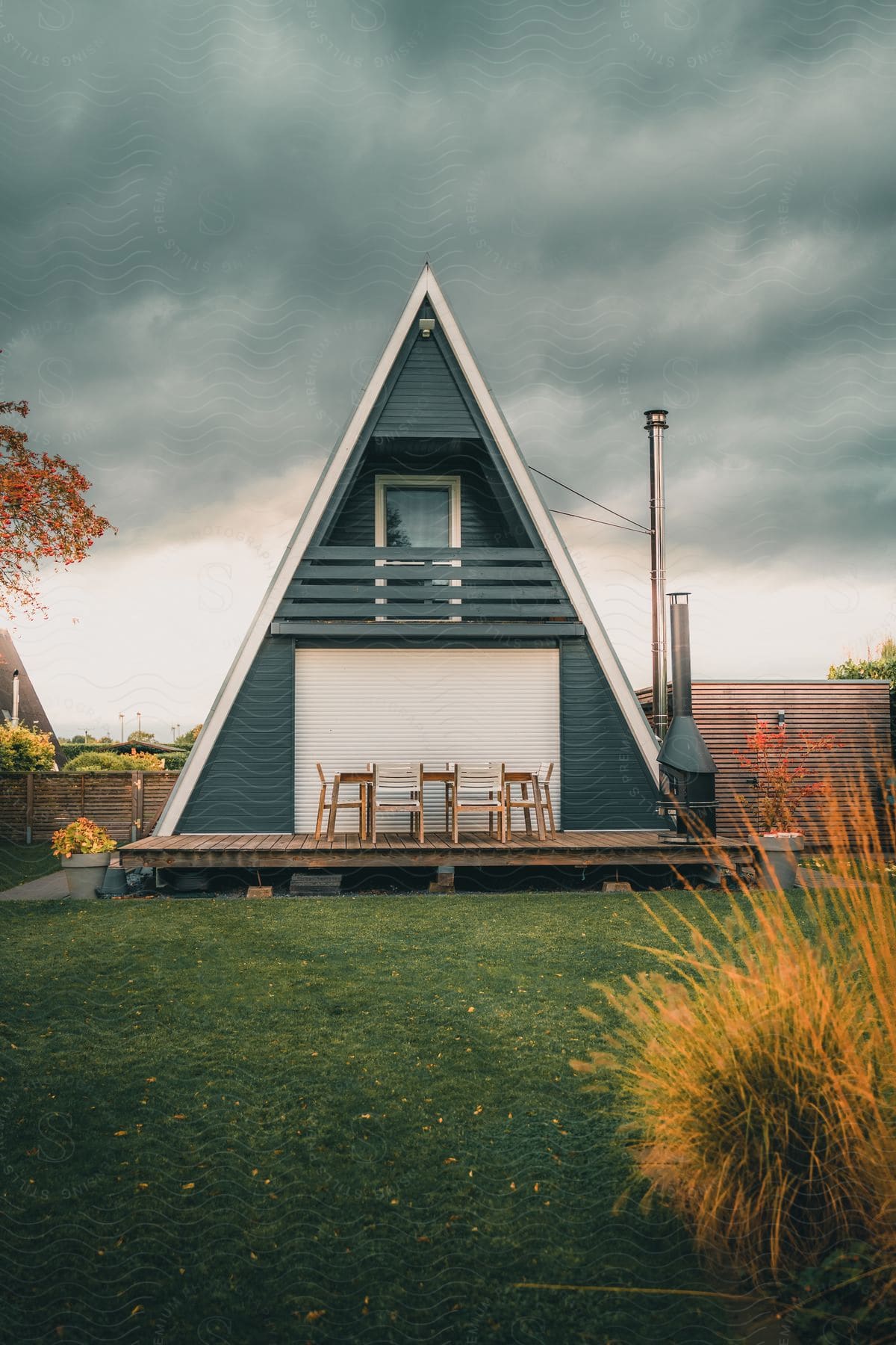 A green triangular house with a deck and colorful trees sits in a yard on a cloudy day.