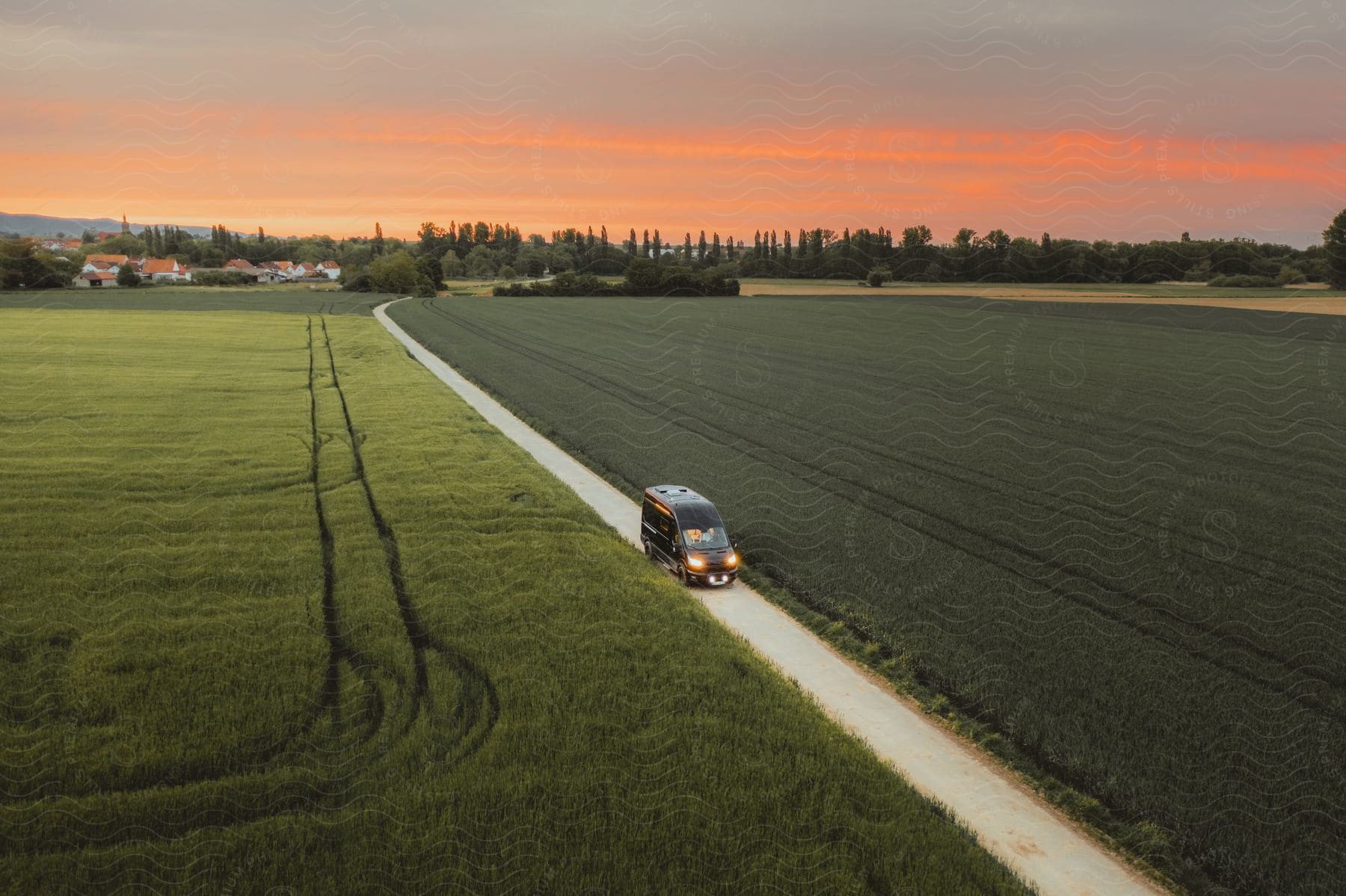 Van driving on country road through green fields at sunset with orange sky.