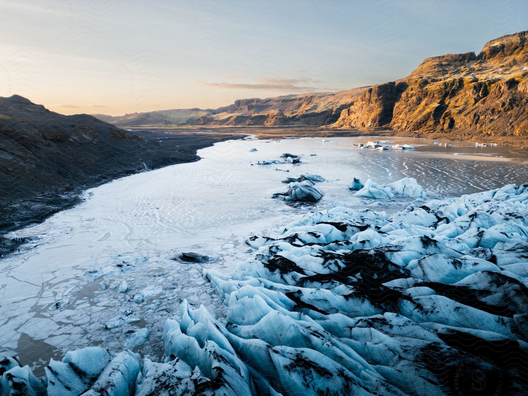 Sun sets over a frozen lake with icebergs, framed by rugged mountains.