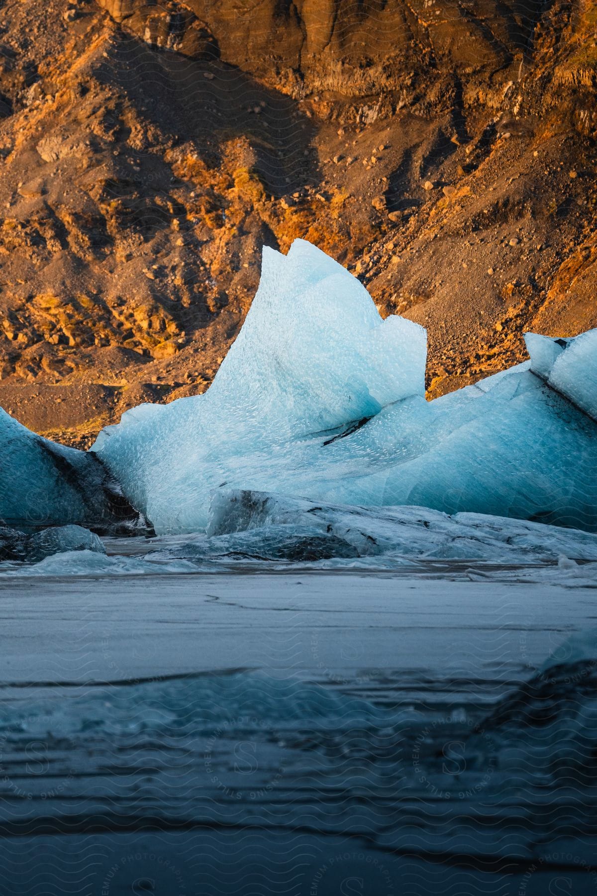 Blue ice formation on water with rocky mountain in the background.