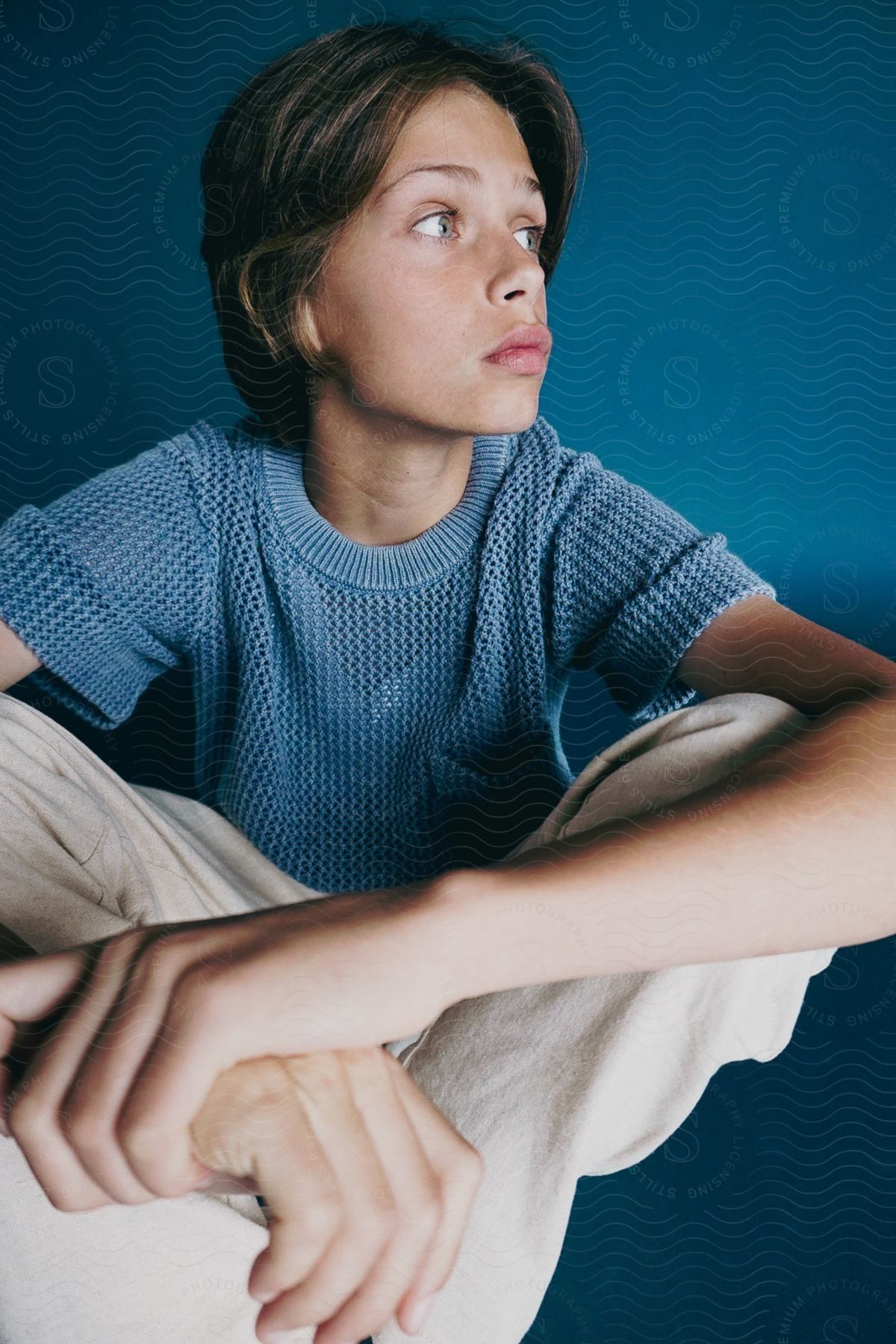 A teenage boy sits against a blue background with his feet crossed.