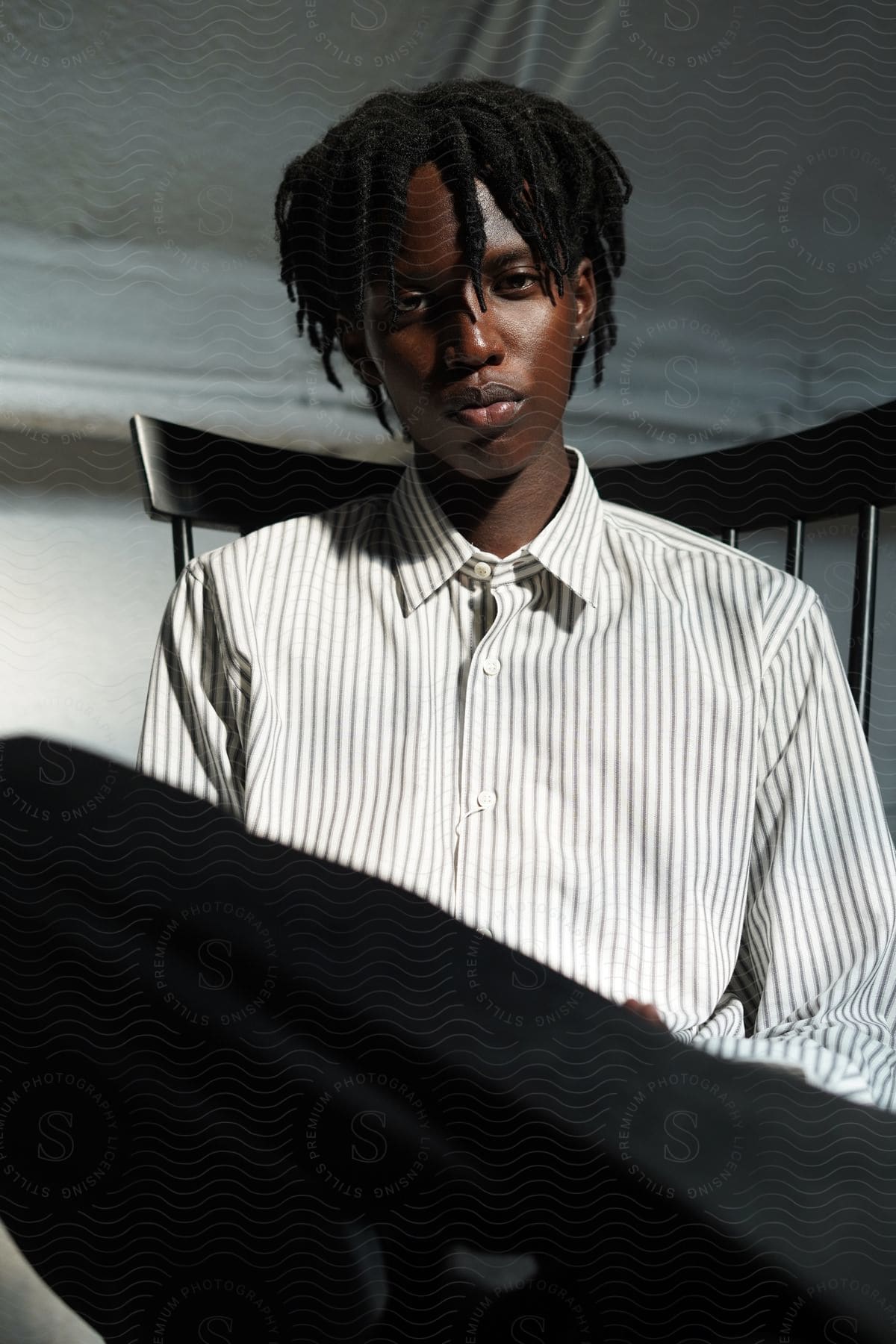 A young man wearing black pants and a striped shirt sits in a chair with his leg up looking sad