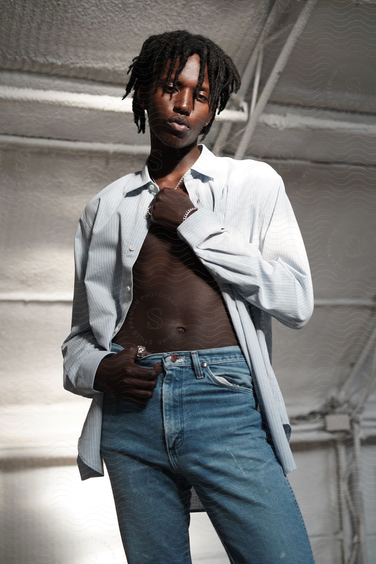 A young man is standing with one hand on top of his jeans and the other hand holding a chain in his open shirt