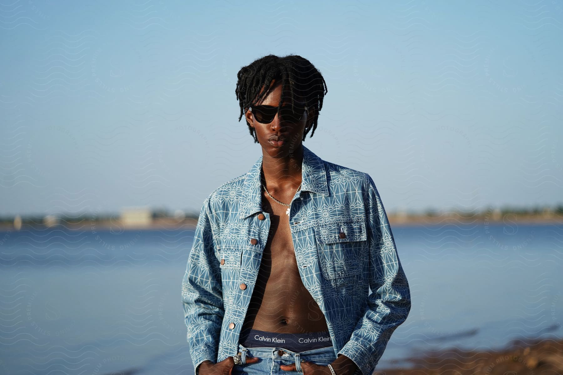 Man in patterned denim jacket and sunglasses by the water at daytime.