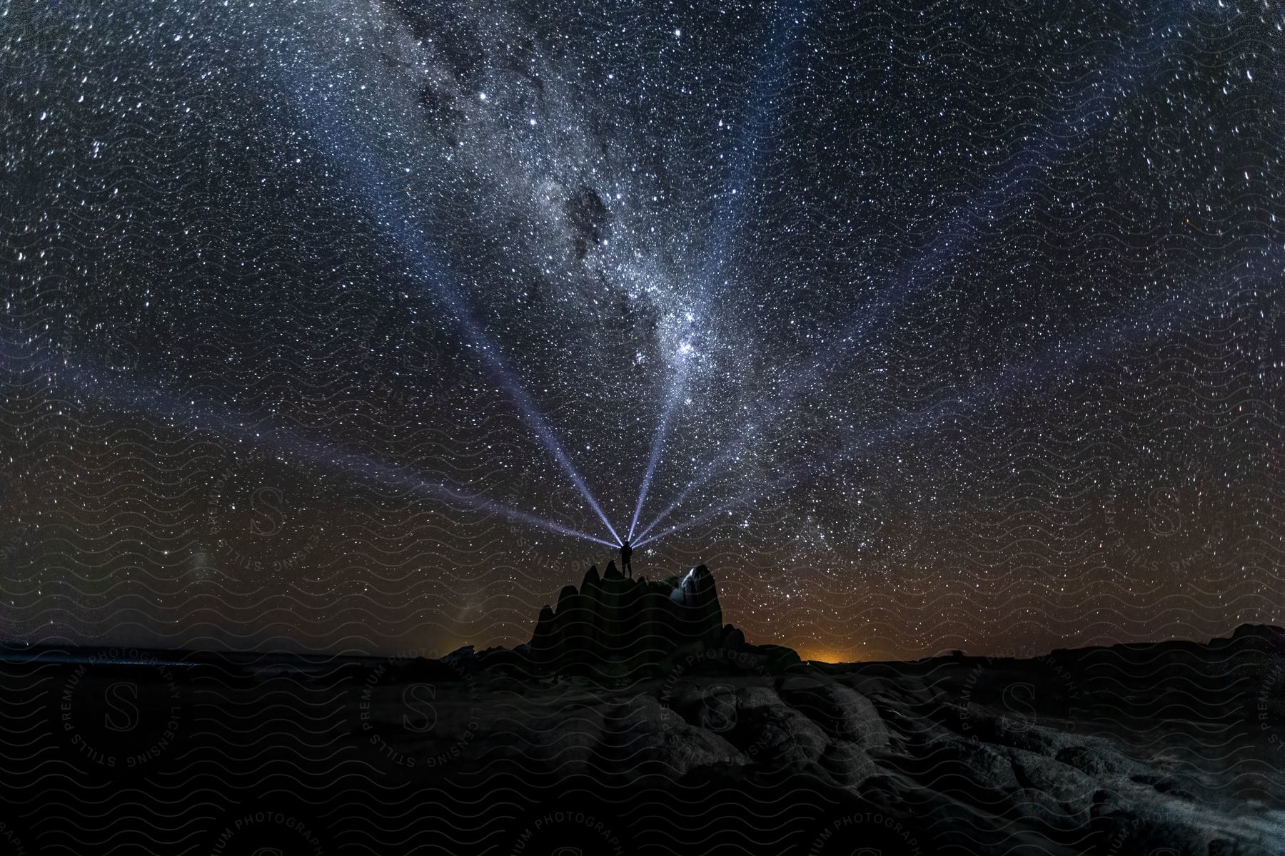 Night sky filled with stars and beams of light radiating from a person standing on a rocky landscape.