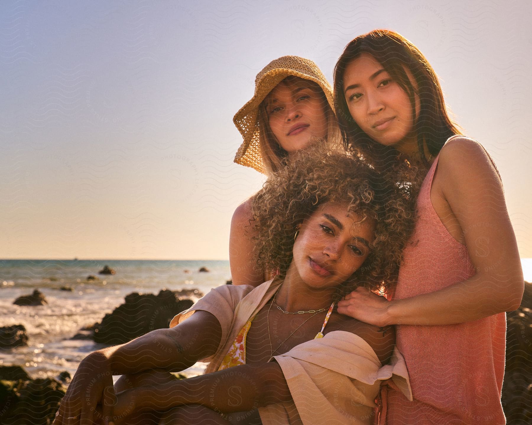 Female friends wearing summer fashions lean together on beach.