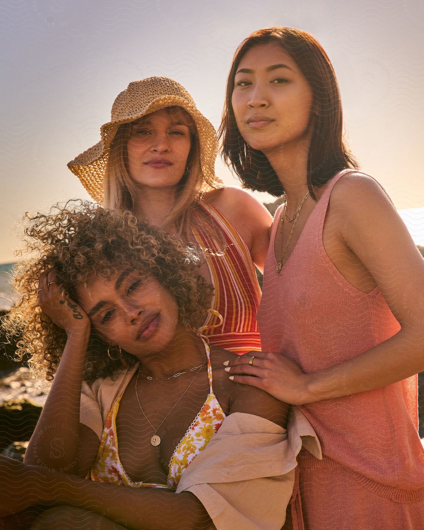 Three women in an outdoor environment looking straight ahead where one of them is sitting and the other two are standing next to and behind her and they are all in an environment close to the sea