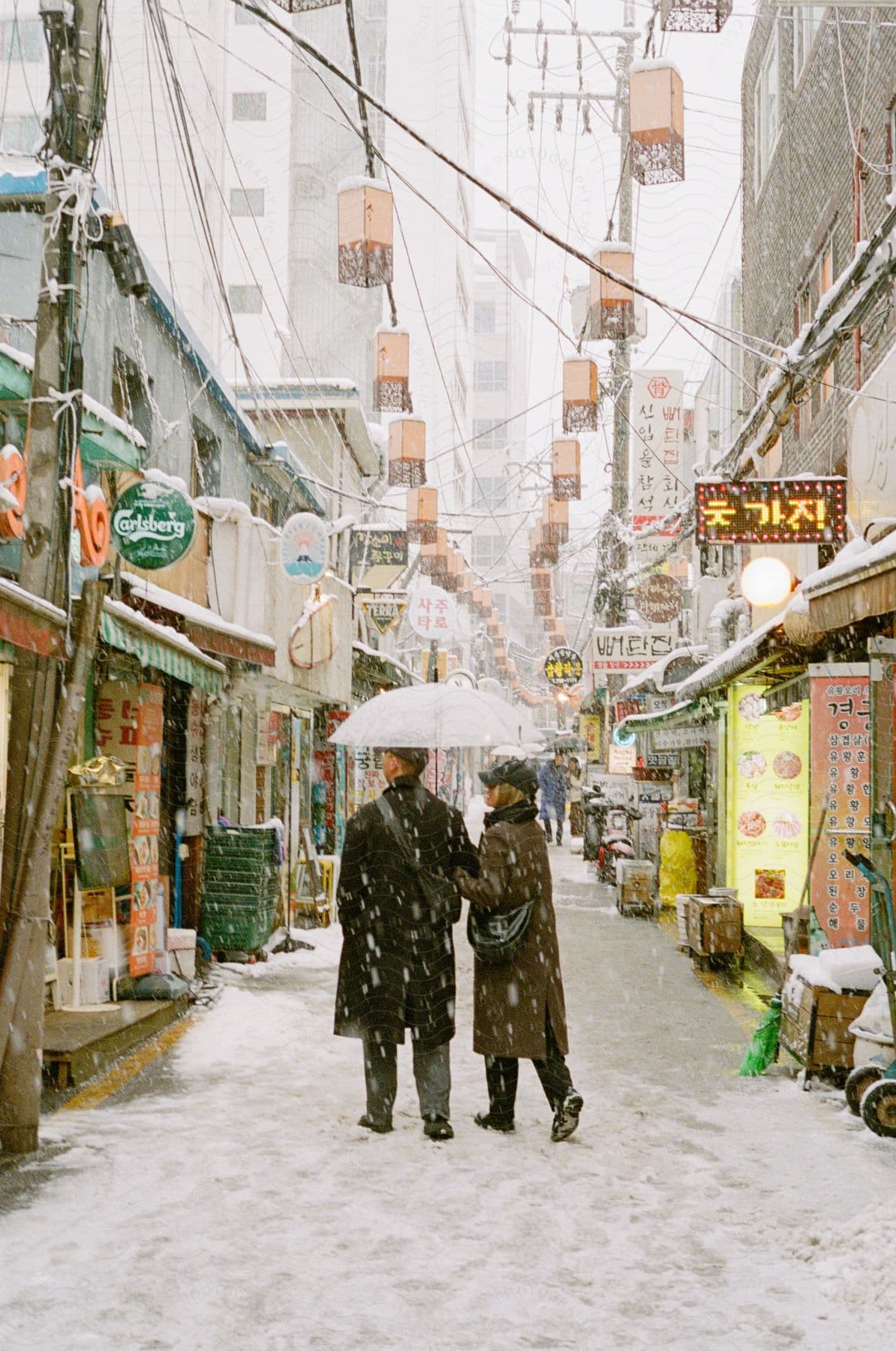 A couple with arms locked and holding an umbrella walks on a snow covered chinese street.