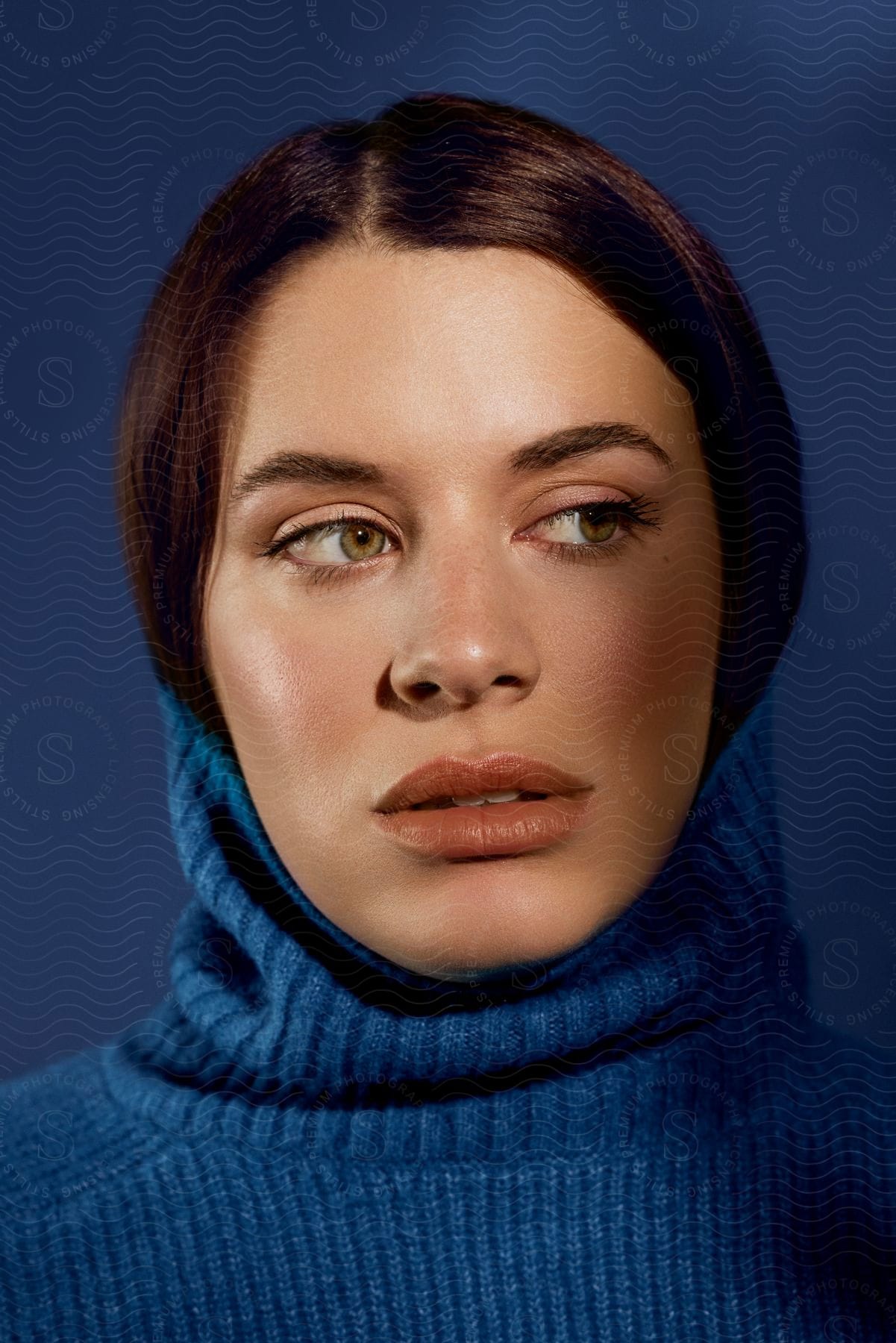 Portrait of a woman with green eyes looking to the side and wearing a blue blouse on a darker blue solid background in contrast to the blouse.