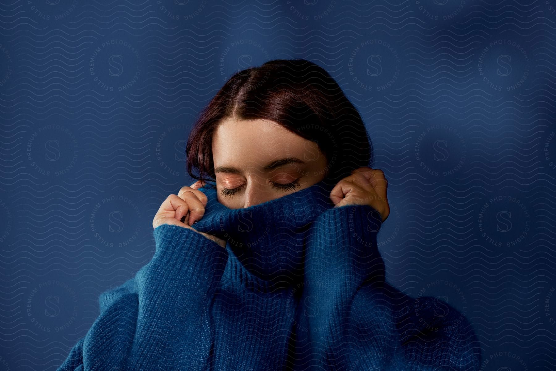 A woman with her eyes closed, wearing a large blue sweater in front of a blue background, pulling the collar up over her nose.