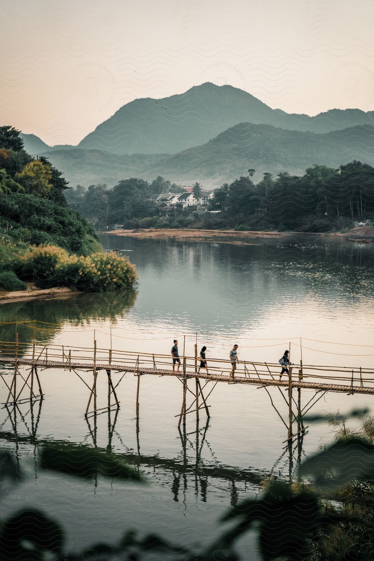 People walk across footbridge a distance from buildings nestled near foggy mountains.