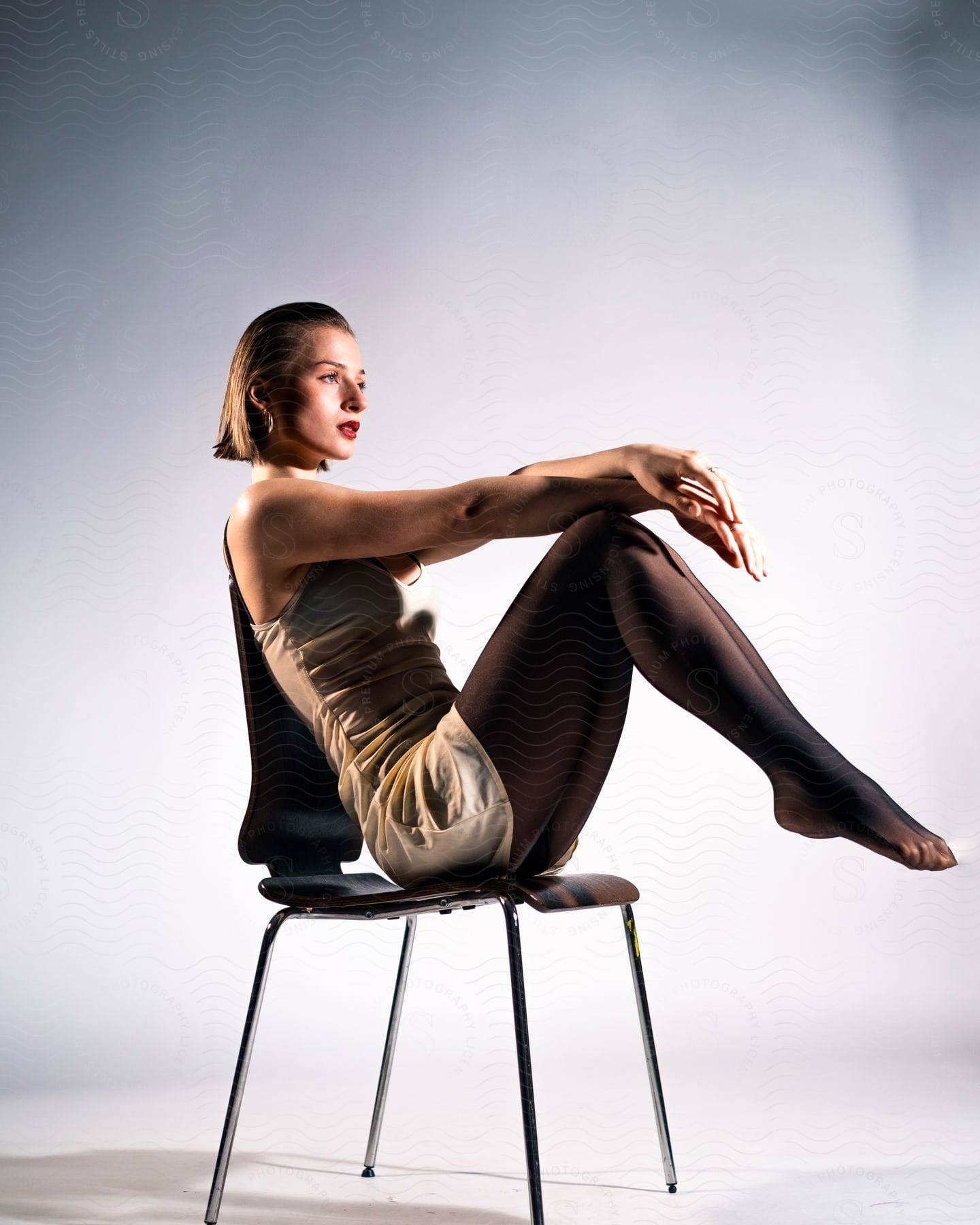 Female with short dark hair, wearing dress, sits on chair with knees elevated, feet off floor