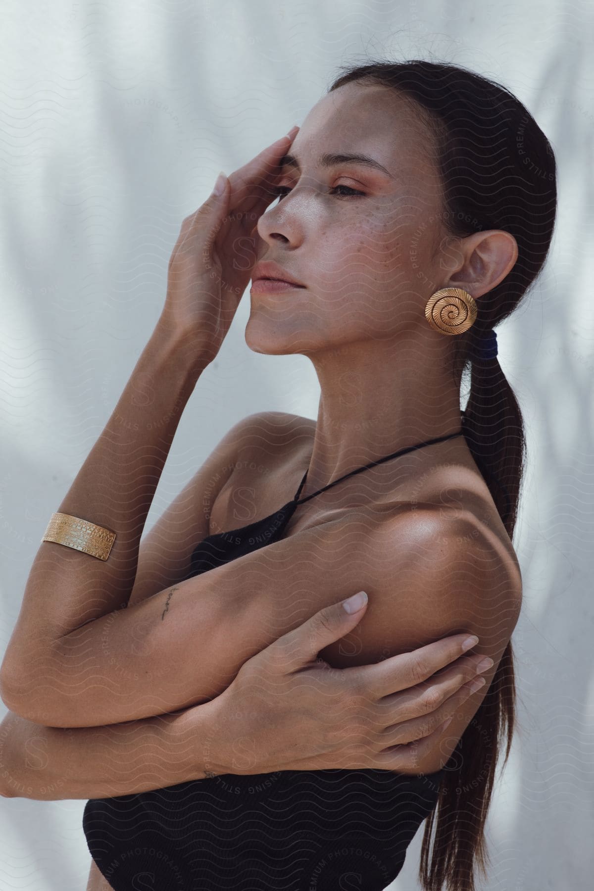 Woman In A Black Sleeveless Top And Gold Cuff Bracelet Touching Her Face And Looking To The Side, With Wears Large Spiral Earrings