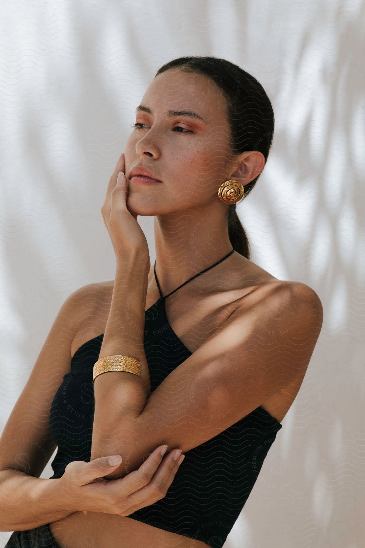 A woman with a ponytail, golden earrings, a bracelet, and a black crop top puts her hand on her cheek.