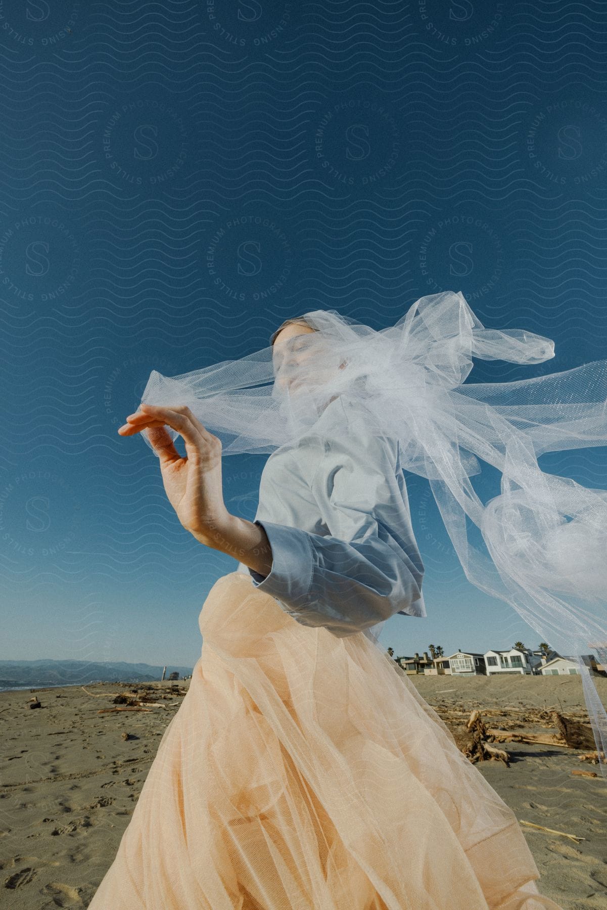 A woman wearing a light blue shirt and pink skirt waves a white veil on the beach near the ocean