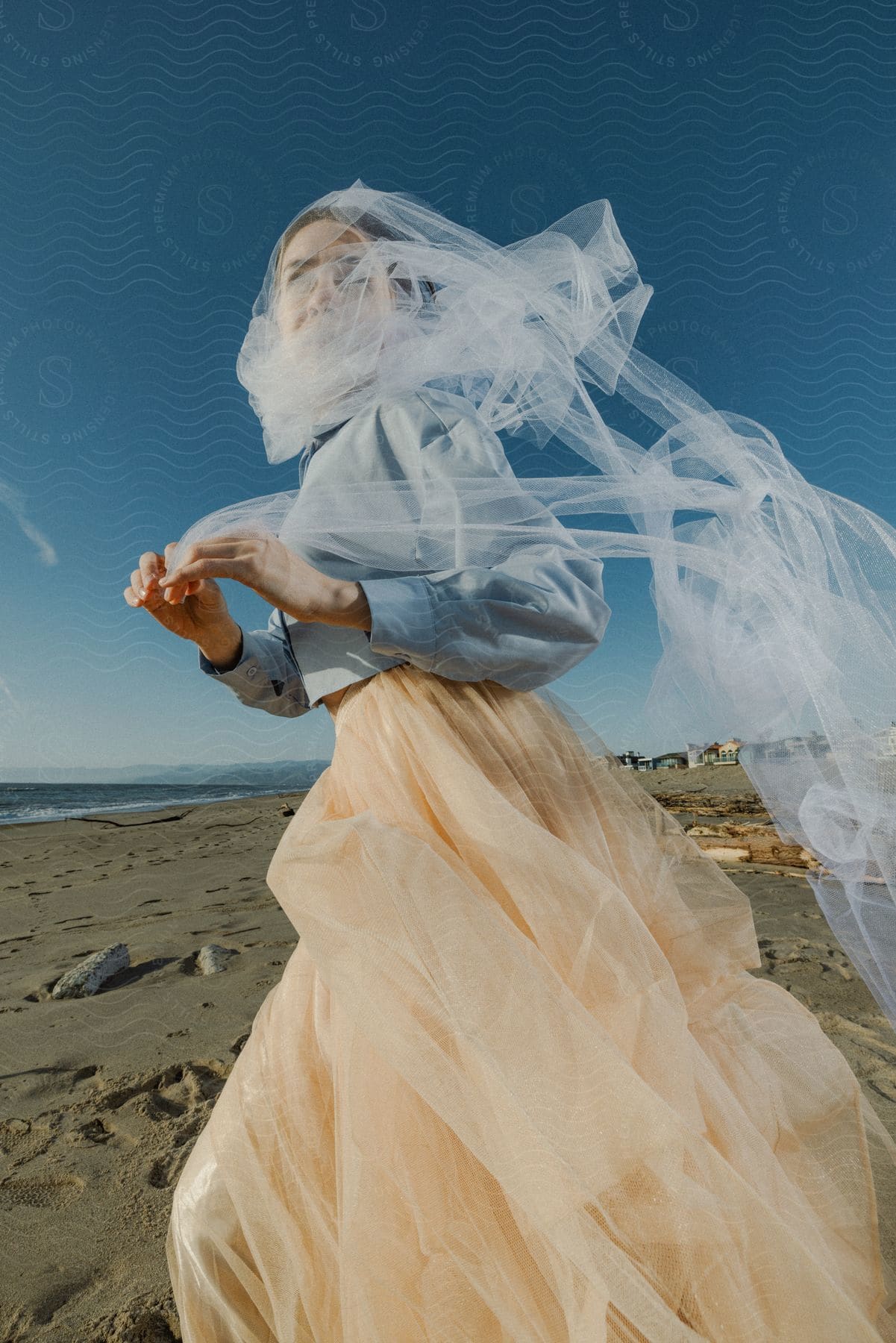 Woman in a blue shirt and peach skirt enveloped in a flowing white veil on a sandy beach against a clear blue sky.