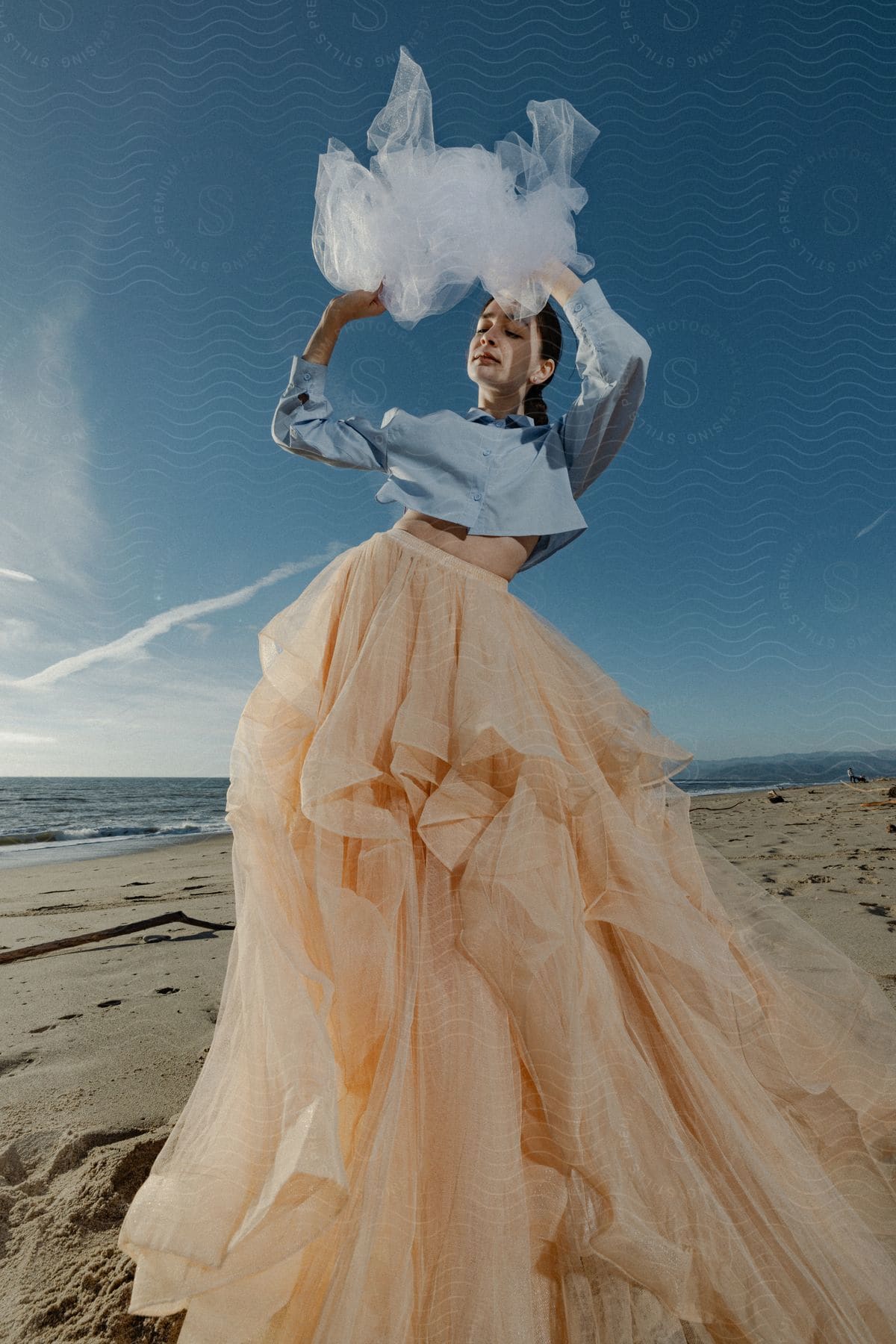 A woman wearing a light blue shirt and pink skirt waves a white veil on the beach.