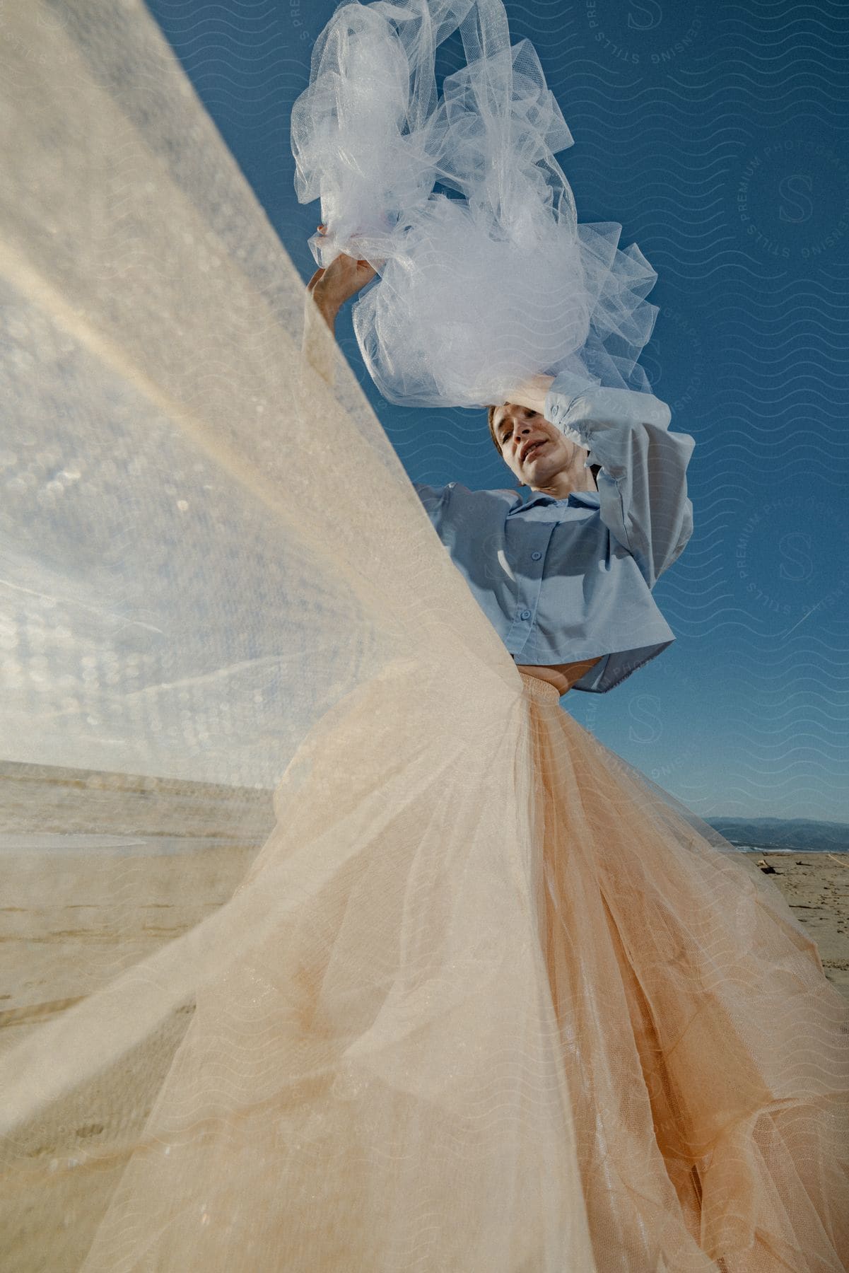 Woman stands on beach in gauzy layered skirt holding a piece of translucent fabric above her head.